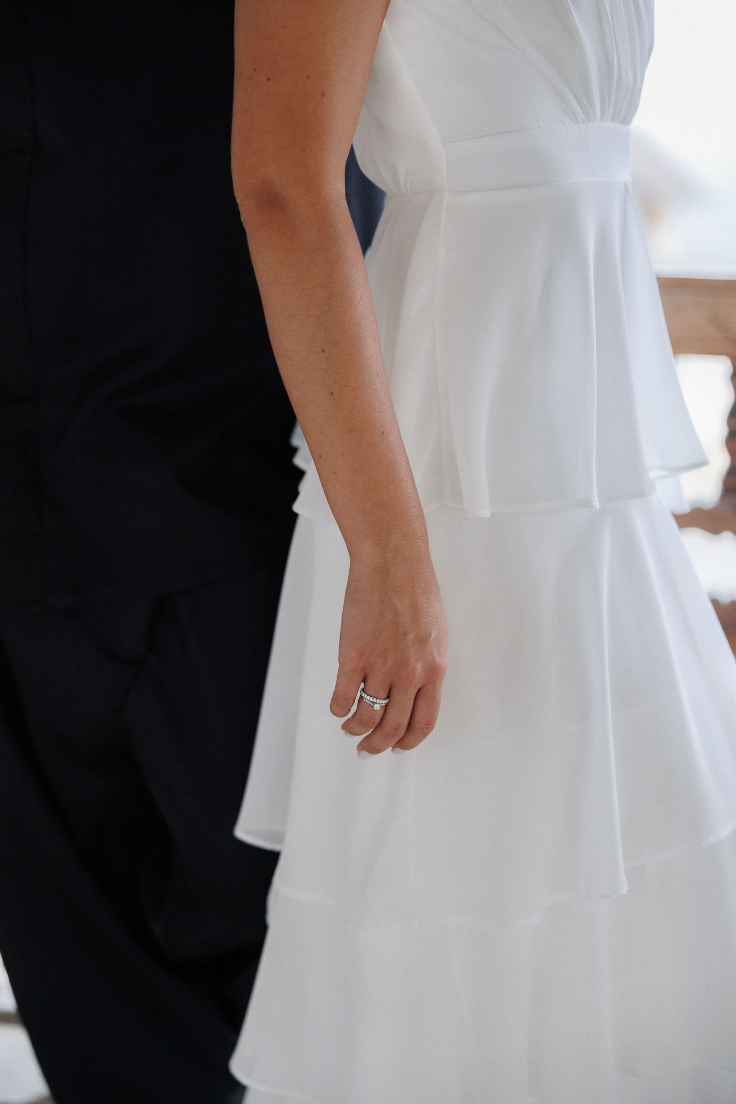 A close-up view of a person wearing a white layered dress standing next to someone in black, with a ring visible on their hand.