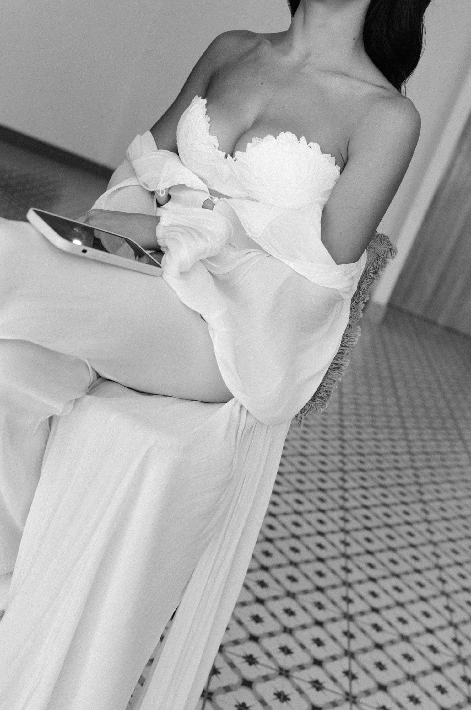 Elegant bride in a strapless white wedding gown seated in a chair, holding a tablet, black and white photo.