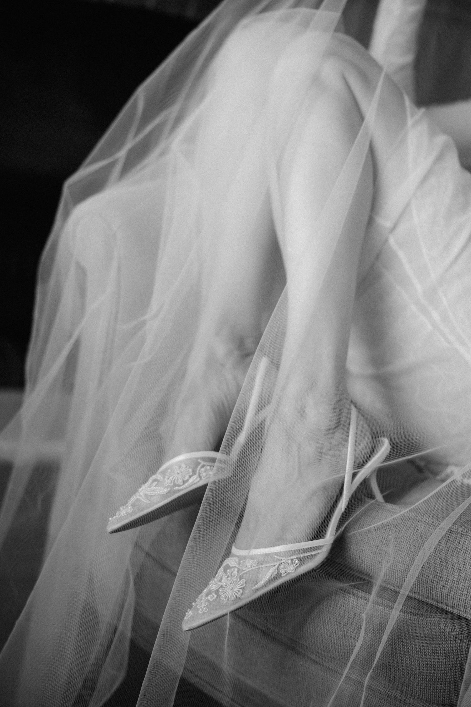Black and white photo of a bride's legs in lace heels beneath a flowing tulle veil.