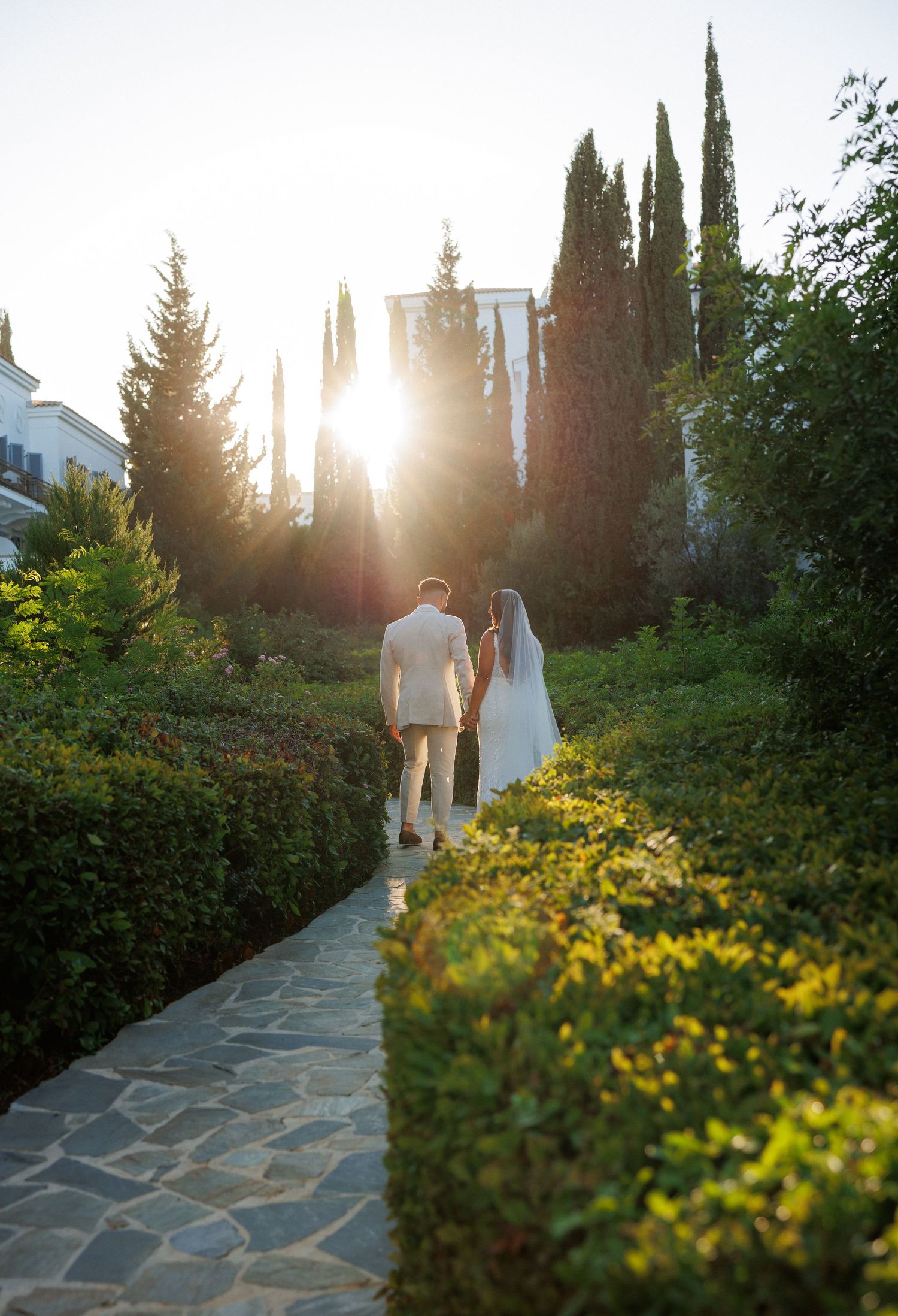 Bride and groom walking hand in hand along a stone garden path at golden sunset.
