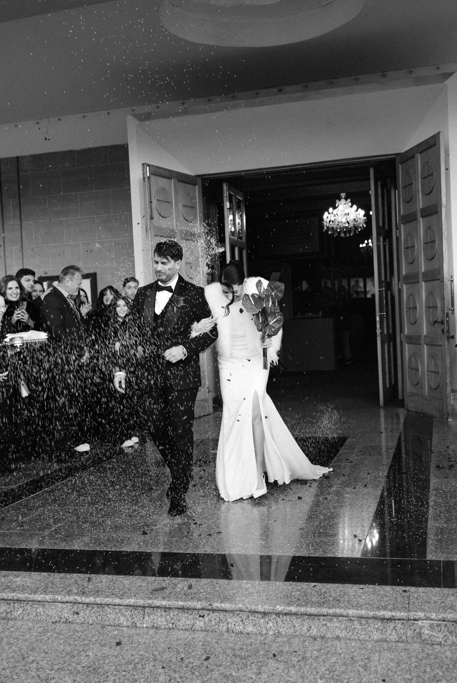 Black and white photo of bride and groom exiting church amid rice shower, guests cheering in background.