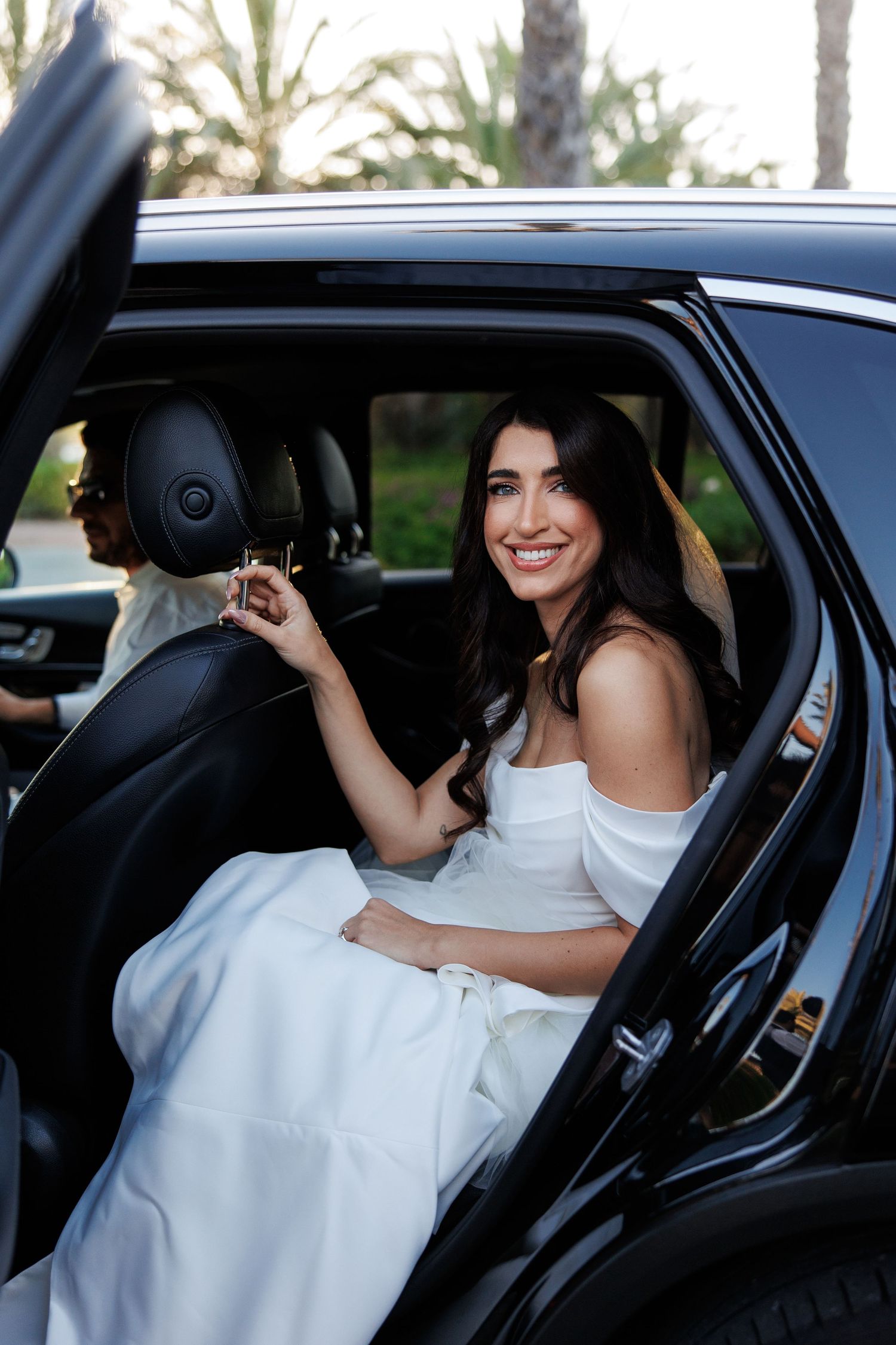 Smiling bride in white off-shoulder gown sitting in a black luxury car, with palm trees visible in the background.