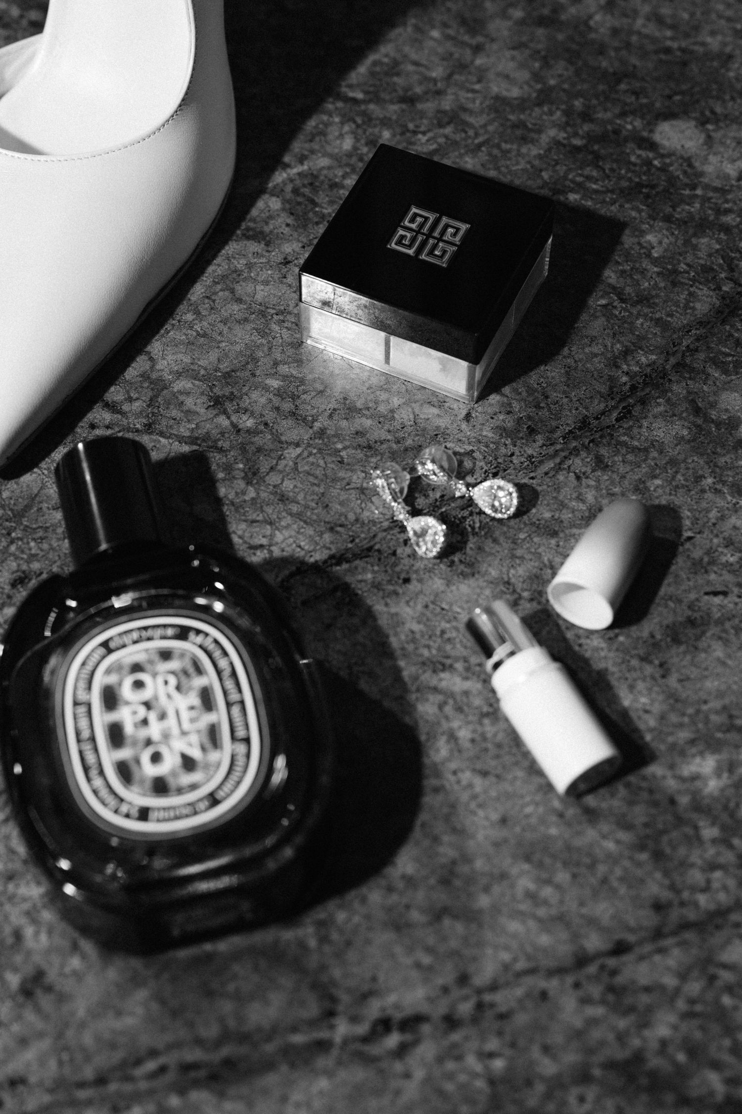 Black and white flatlay of Diptyque perfume, Givenchy compact, lipstick, and earrings on marble surface.