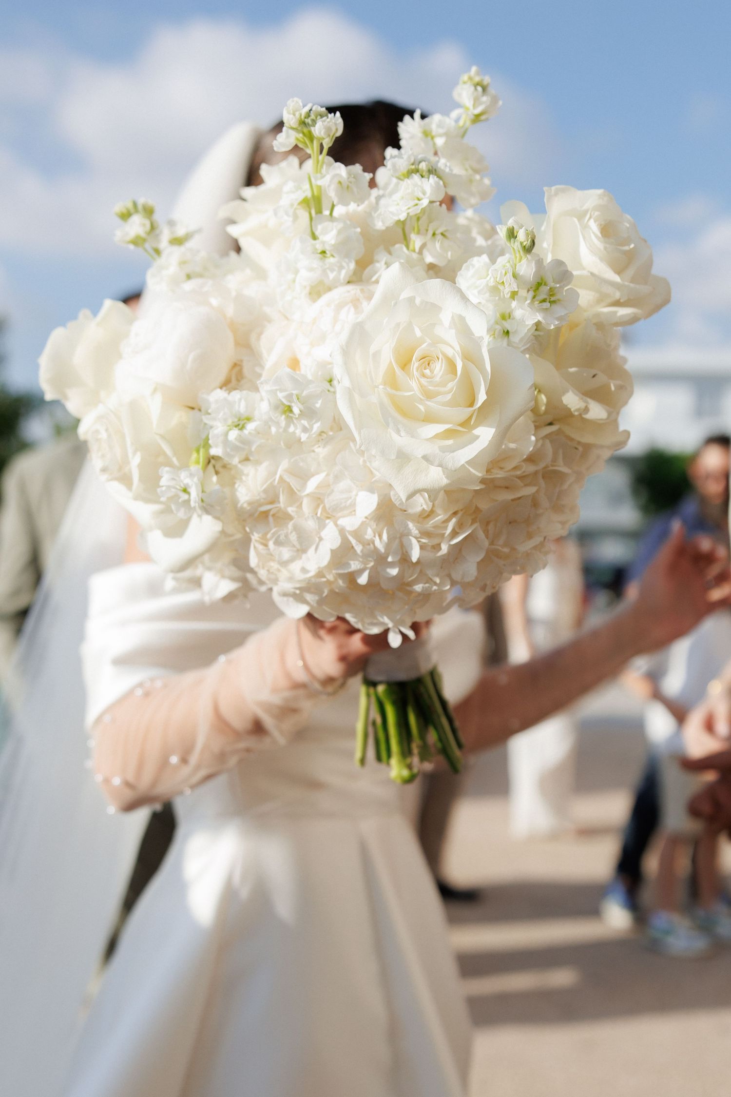 Bride in white wedding dress holding elegant bouquet of white roses and hydrangeas outdoors on sunny day.