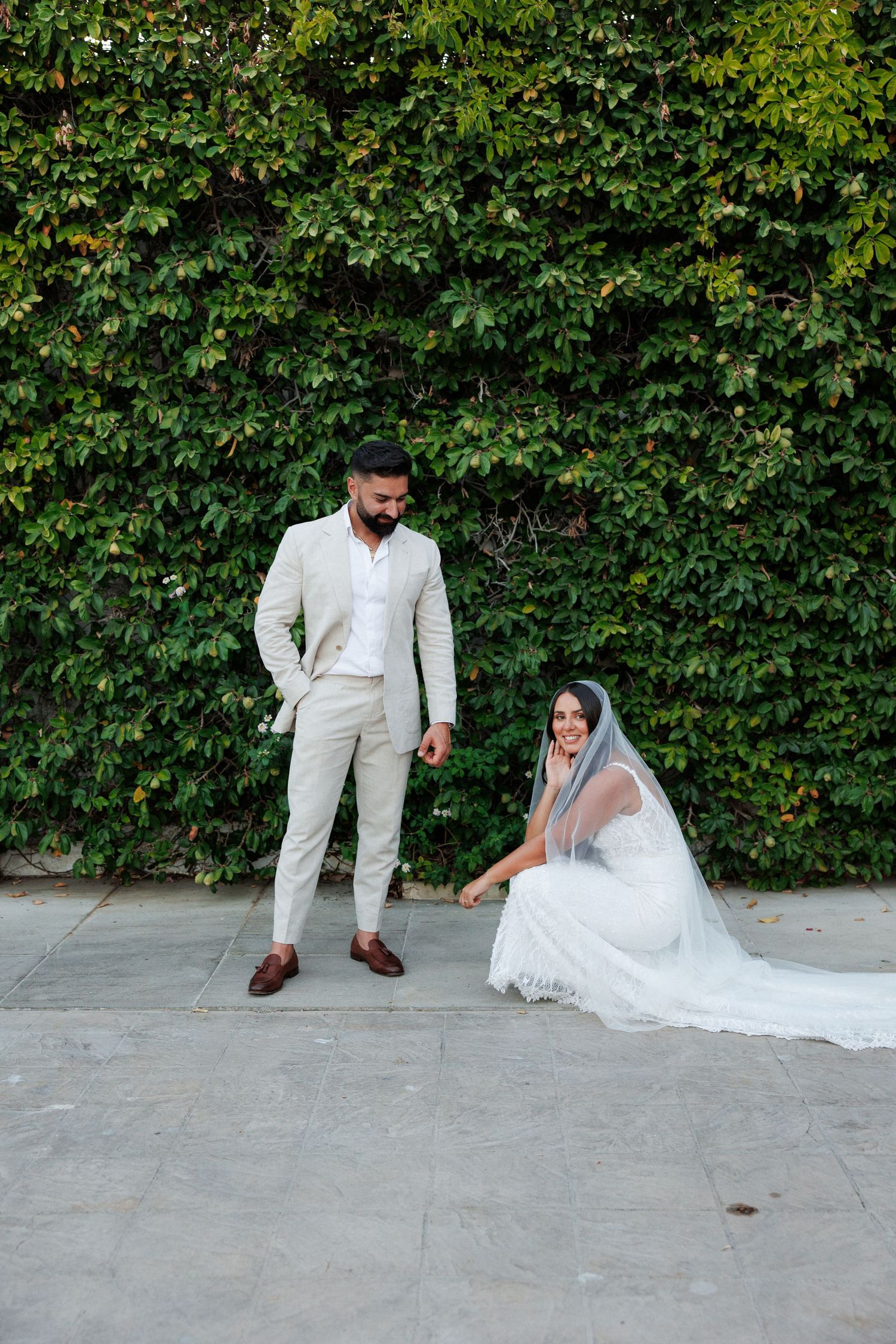 Bride sitting on ground smiling up at groom standing before a lush green ivy wall backdrop.