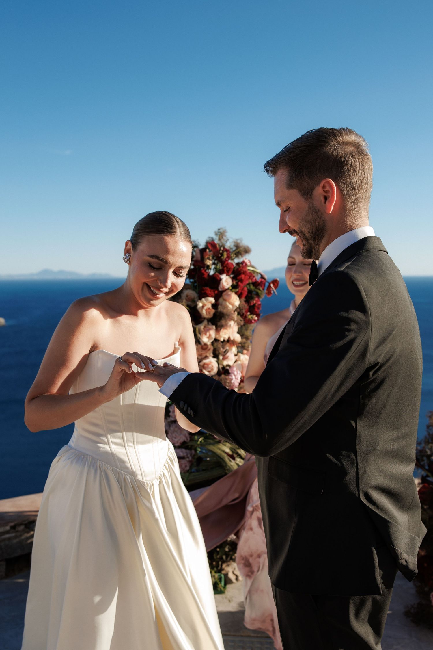 Bride smiles as groom places wedding ring on her finger at outdoor ceremony with ocean view in Santorini.