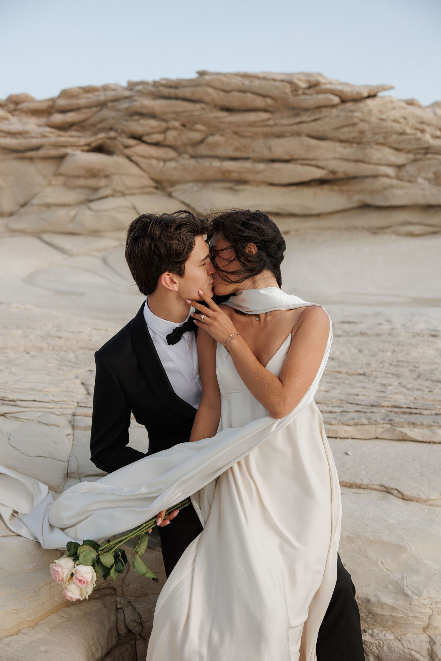 A couple in wedding attire embrace against a backdrop of white stone cliffs and desert landscape.