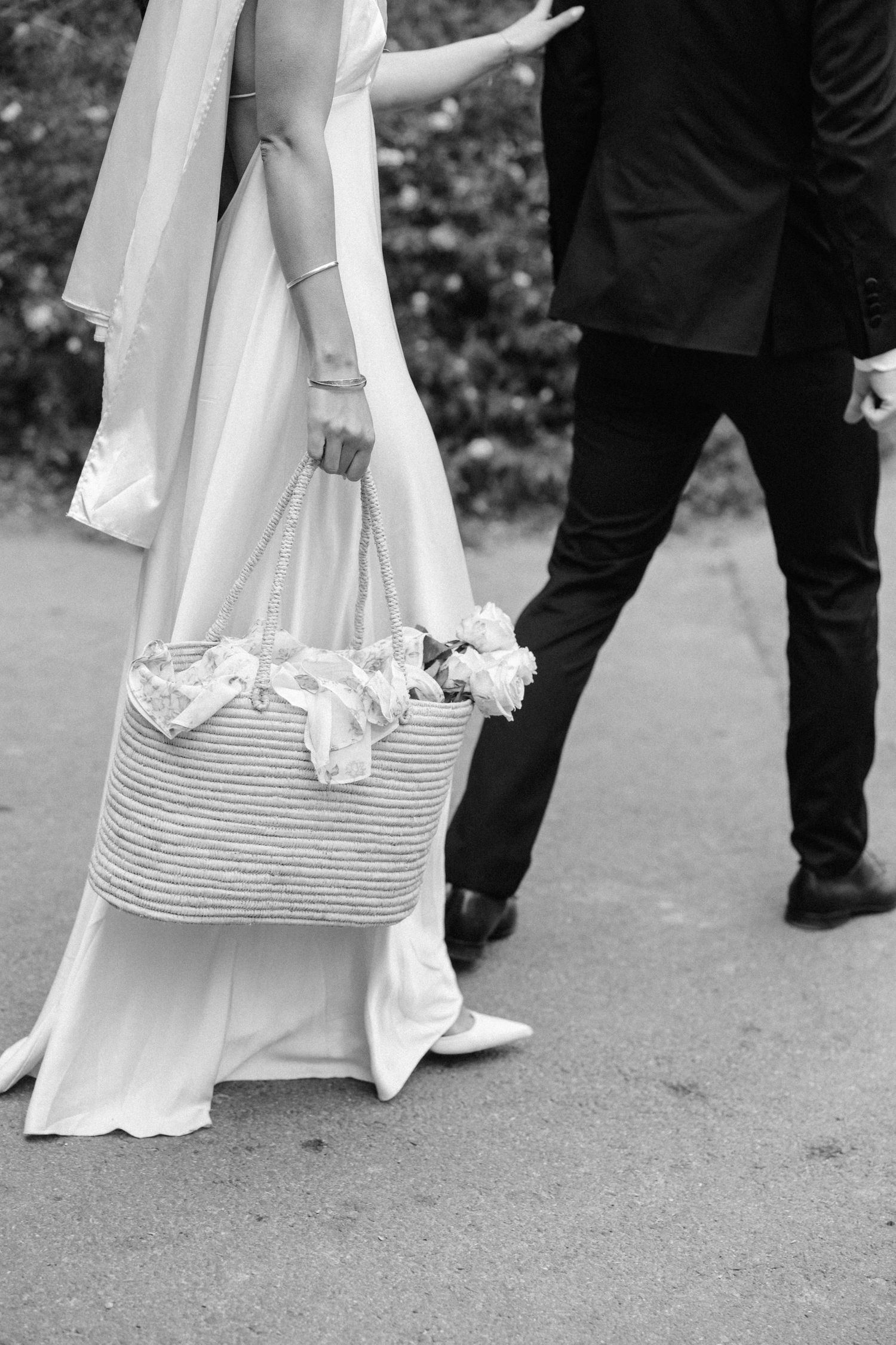 Black and white detail shot of bride with straw bag and groom in formal attire.
