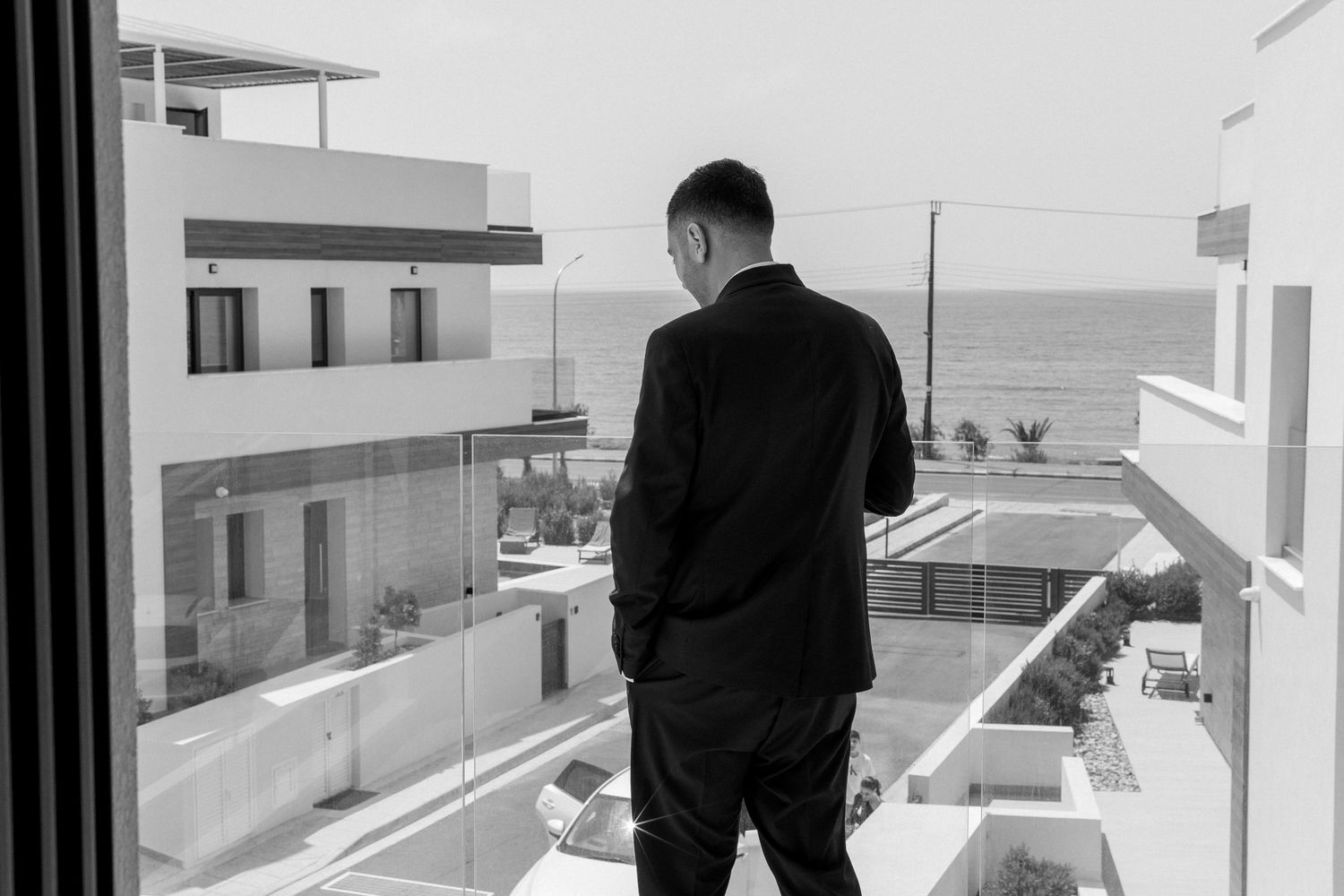 A person in black attire stands on a modern balcony overlooking the ocean and white buildings below.