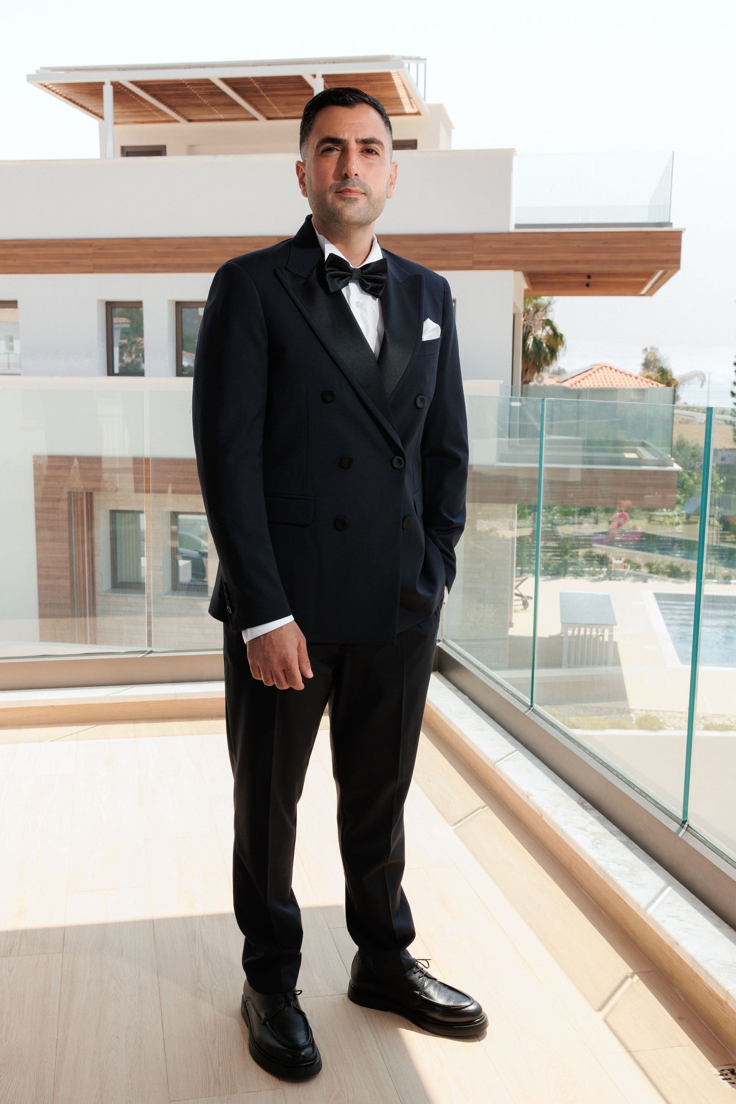 Someone wearing a formal black tuxedo and bowtie stands in a modern balcony space with glass railings and wooden accents.