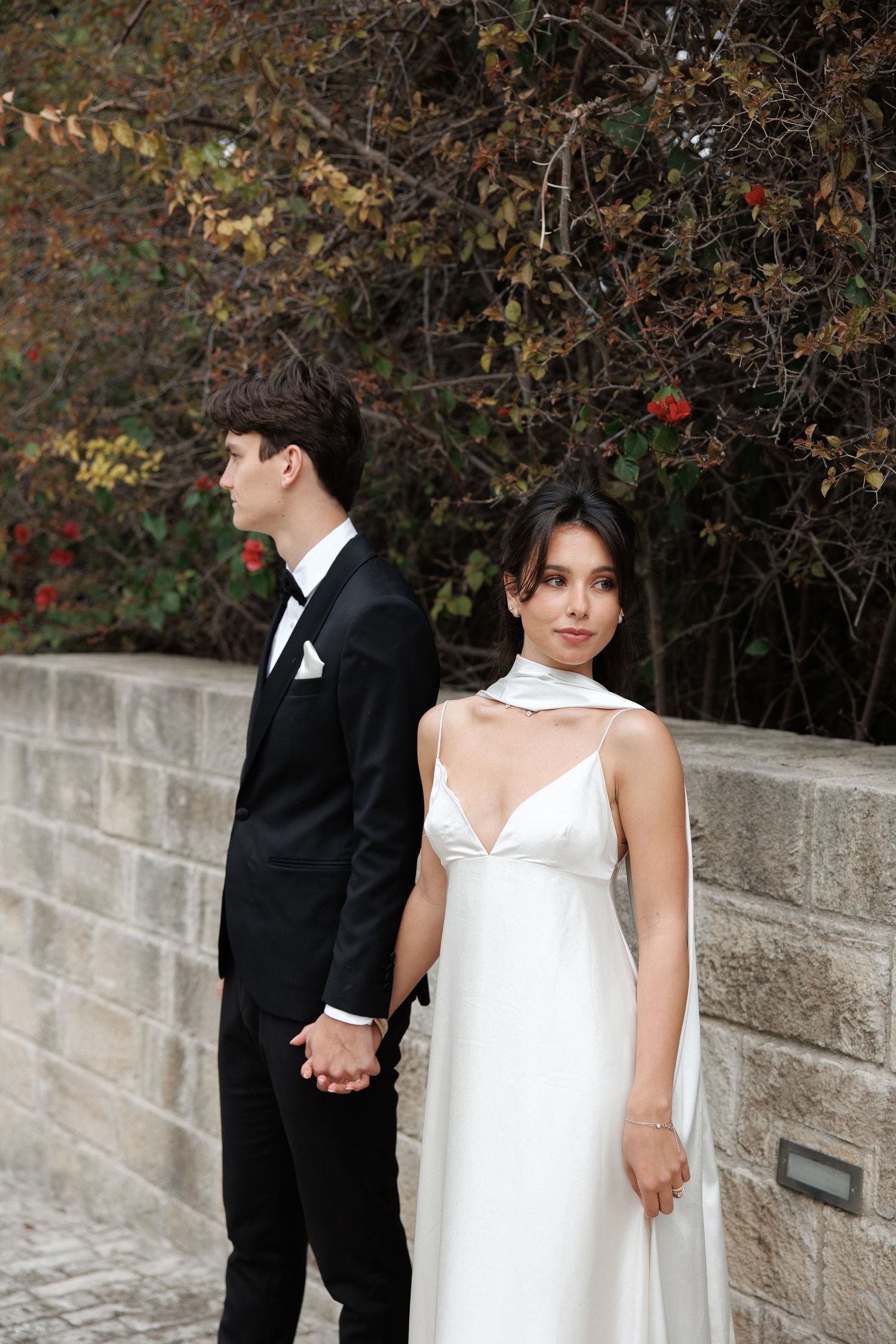 A couple in formal wedding attire holding hands against a stone wall with autumn foliage in the background.