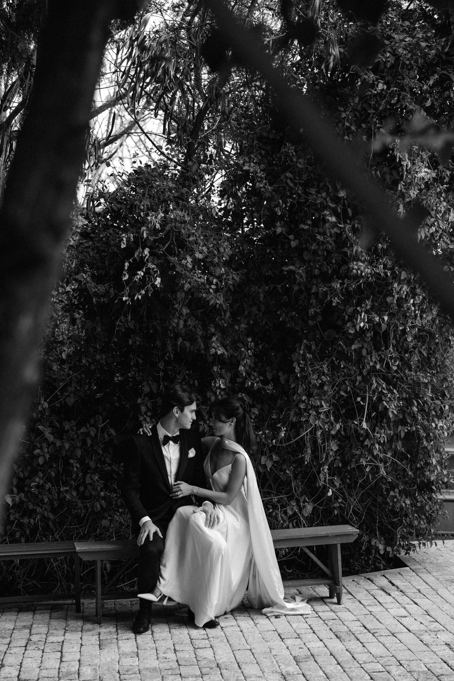 A black and white wedding photo capturing a tender moment between newlyweds seated on a park bench under trees.