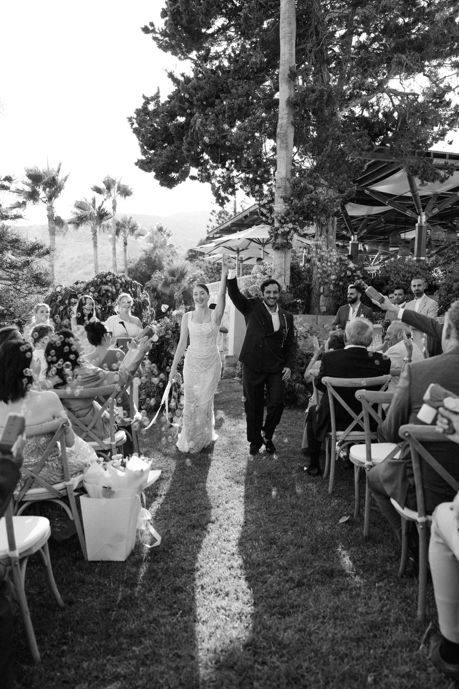 Newlyweds celebrate recessional with raised hands as guests cheer in black and white outdoor wedding photo.
