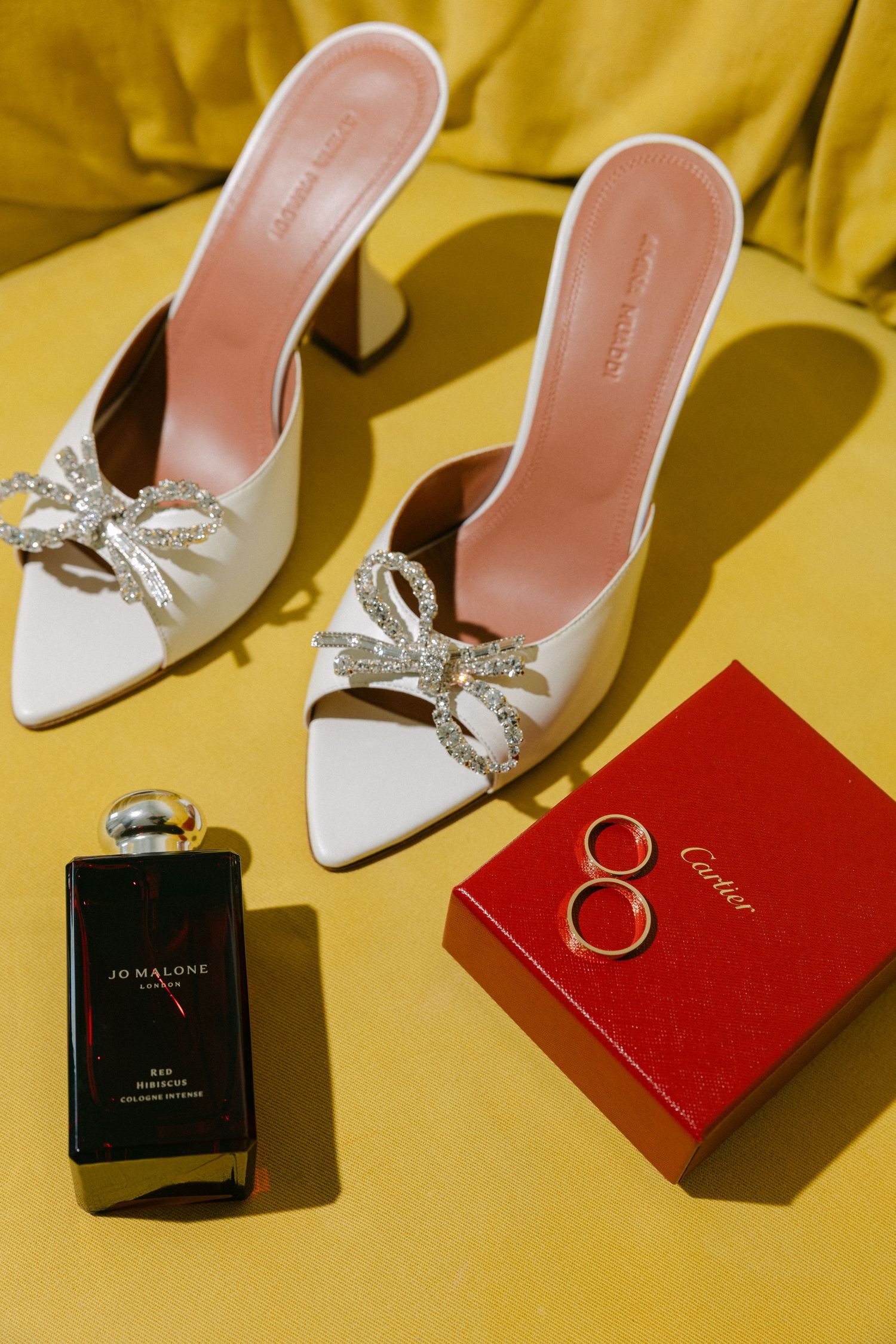 Elegant white bridal shoes with crystal embellishments and perfume bottles arranged on yellow background.