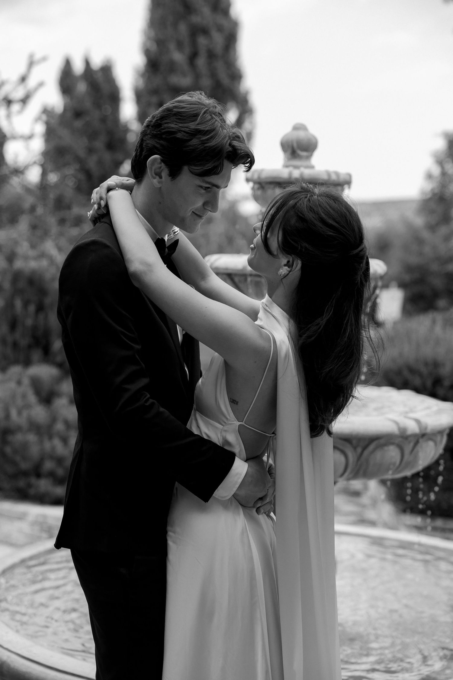 A romantic black and white photo of a couple embracing by a garden fountain during an elegant outdoor wedding.
