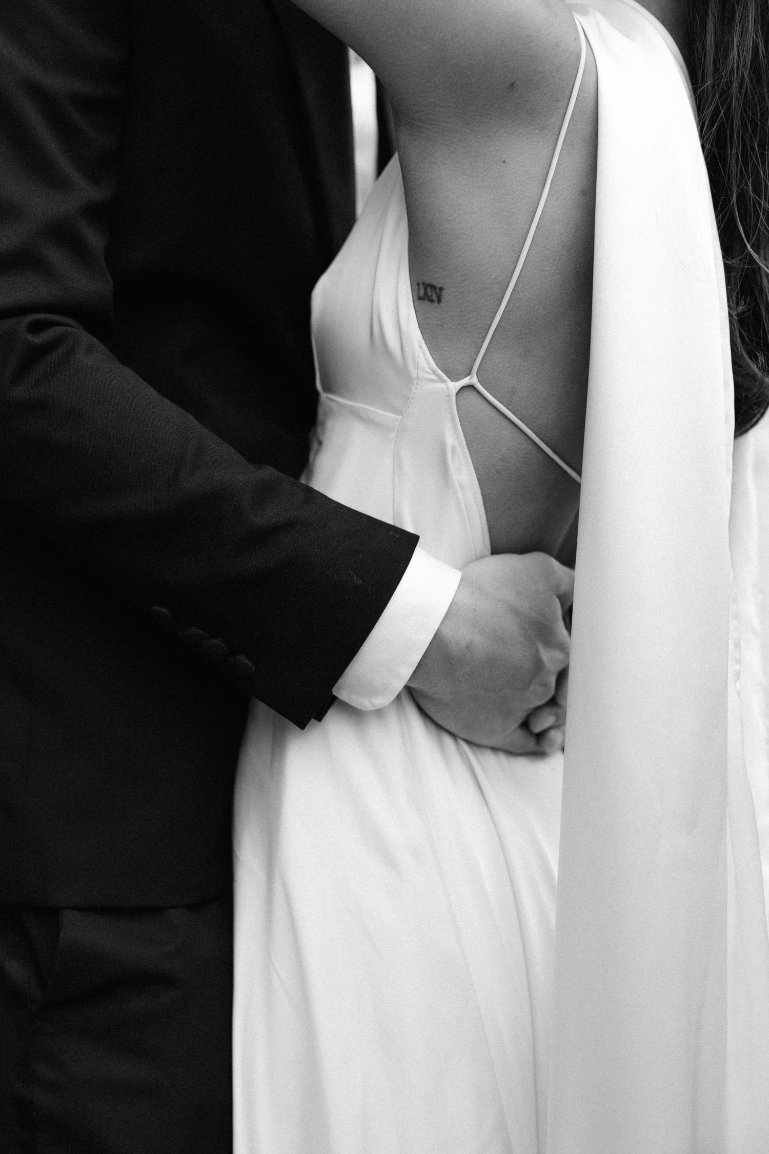 Black and white intimate wedding photo showing hands embracing waist of bride in elegant white dress.