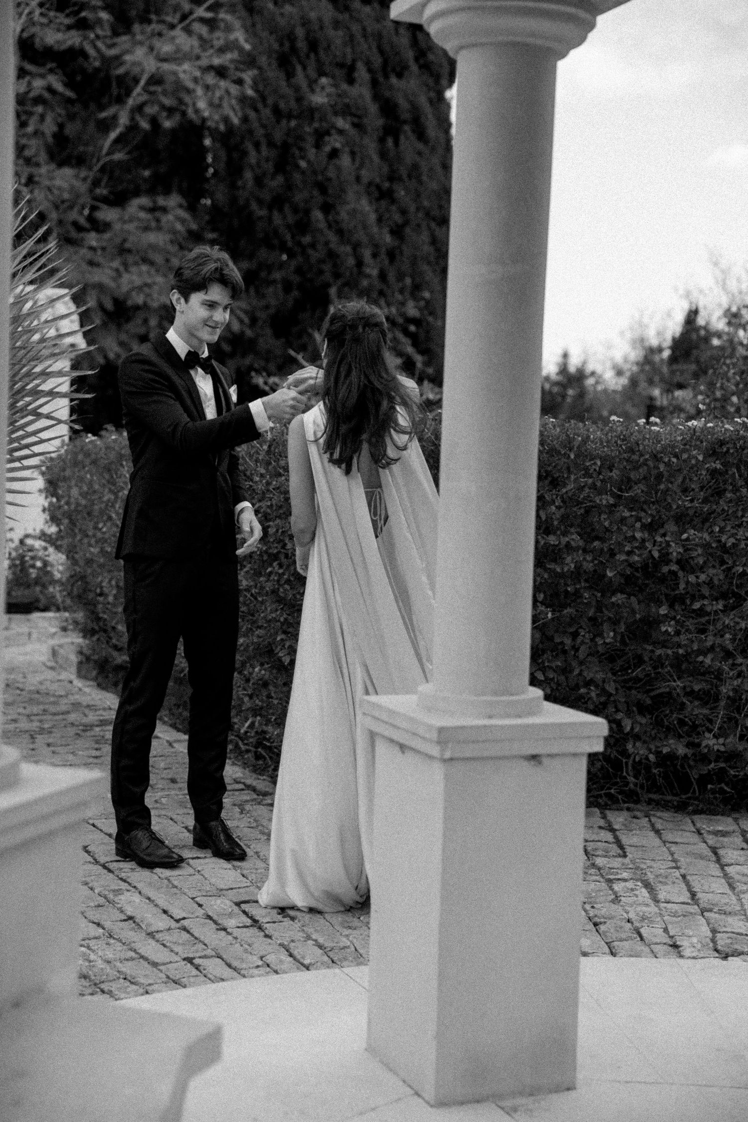 A romantic black and white wedding scene with a couple dancing by a classical column on a stone patio.