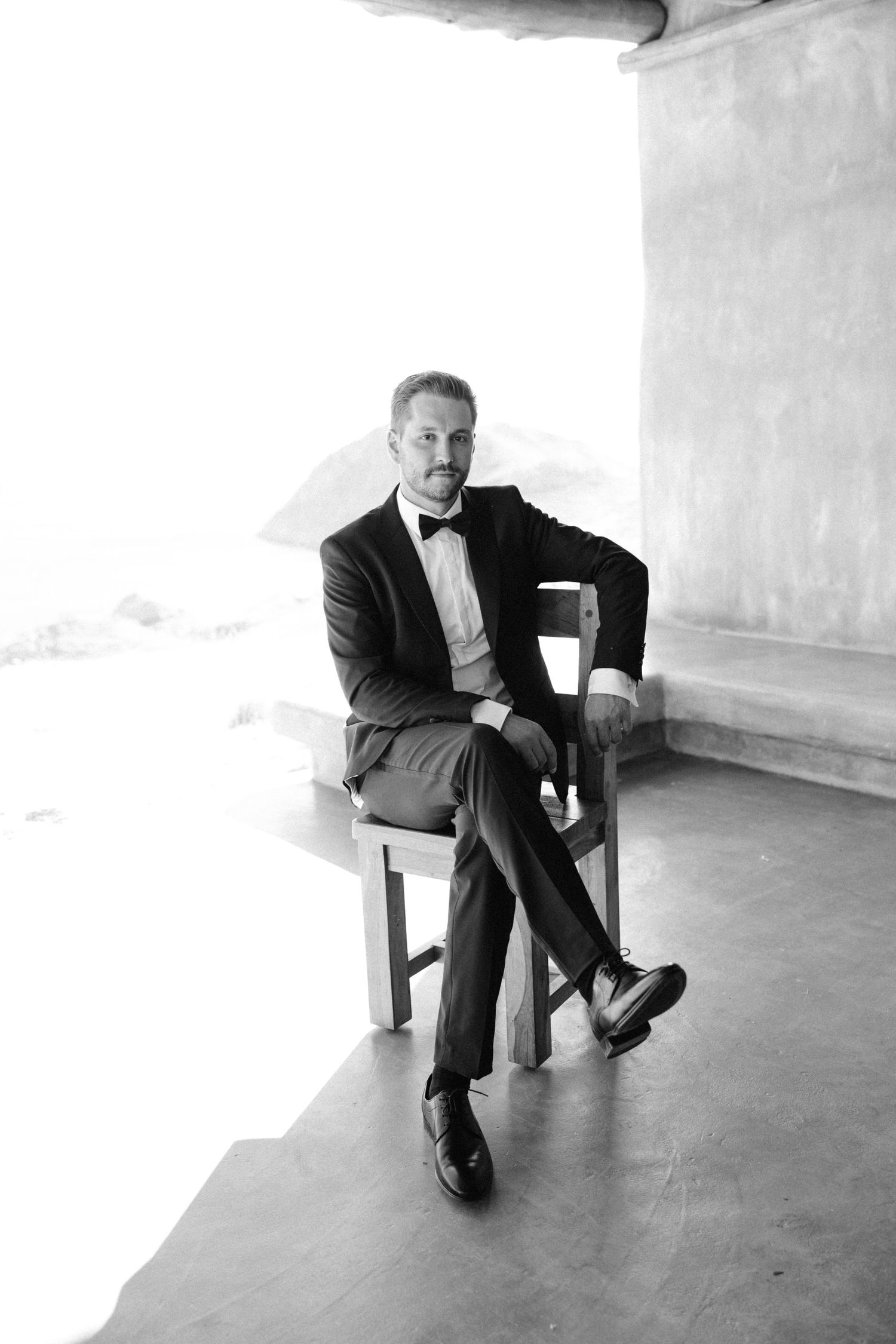 Elegant man in tuxedo with bow tie sitting cross-legged on wooden chair in black and white portrait photo.