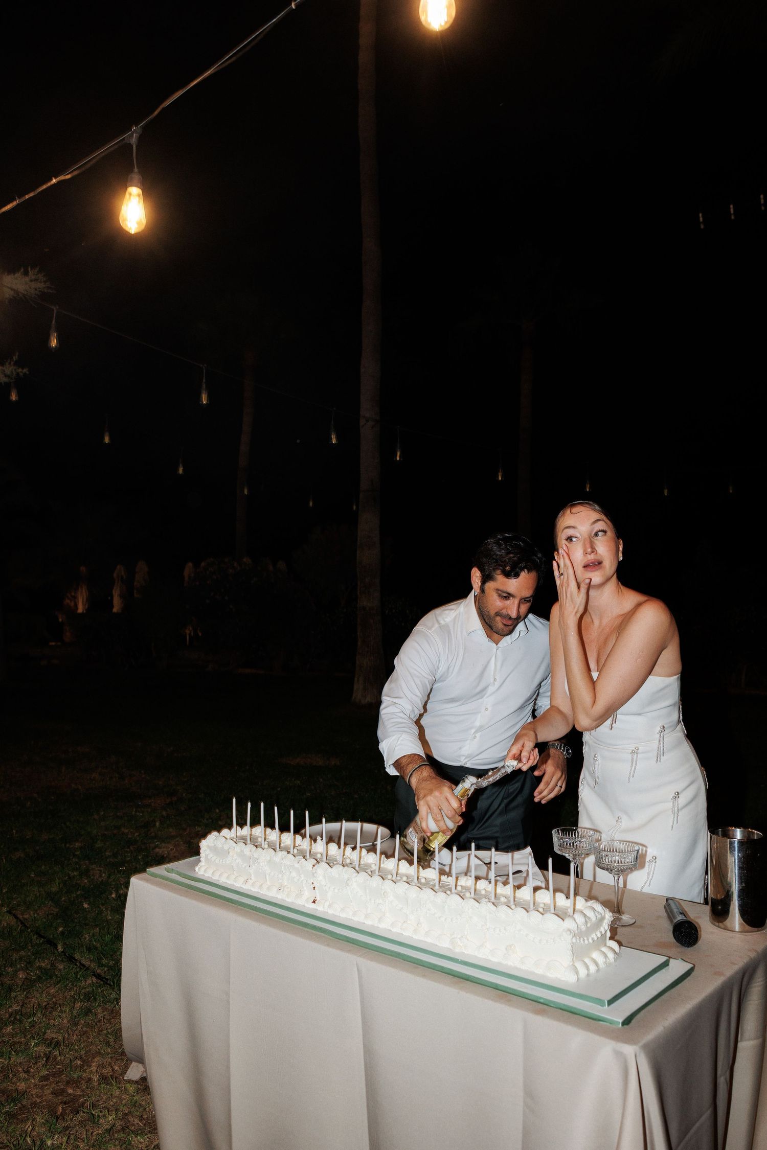 Bride and groom cutting their white wedding cake outdoors at night under warm string lights.