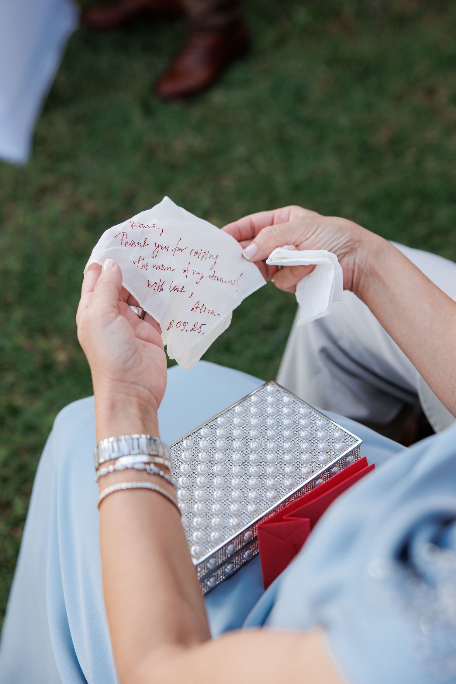 Guest reads handwritten thank you note while holding silver clutch purse at outdoor wedding ceremony.