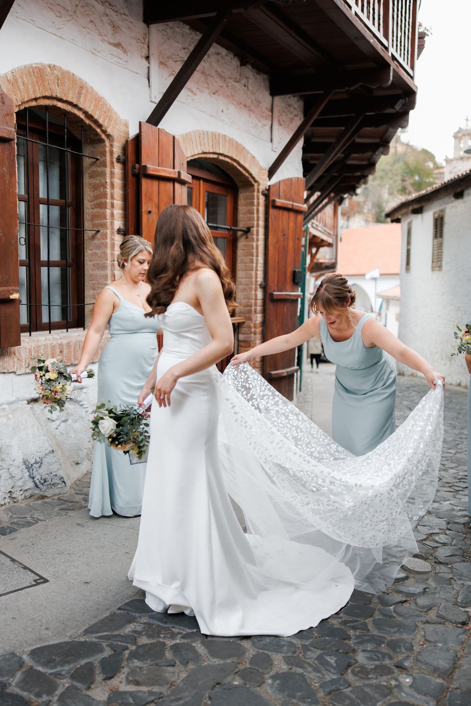 Bridesmaids help arrange the train of a lace wedding dress on cobblestone street.