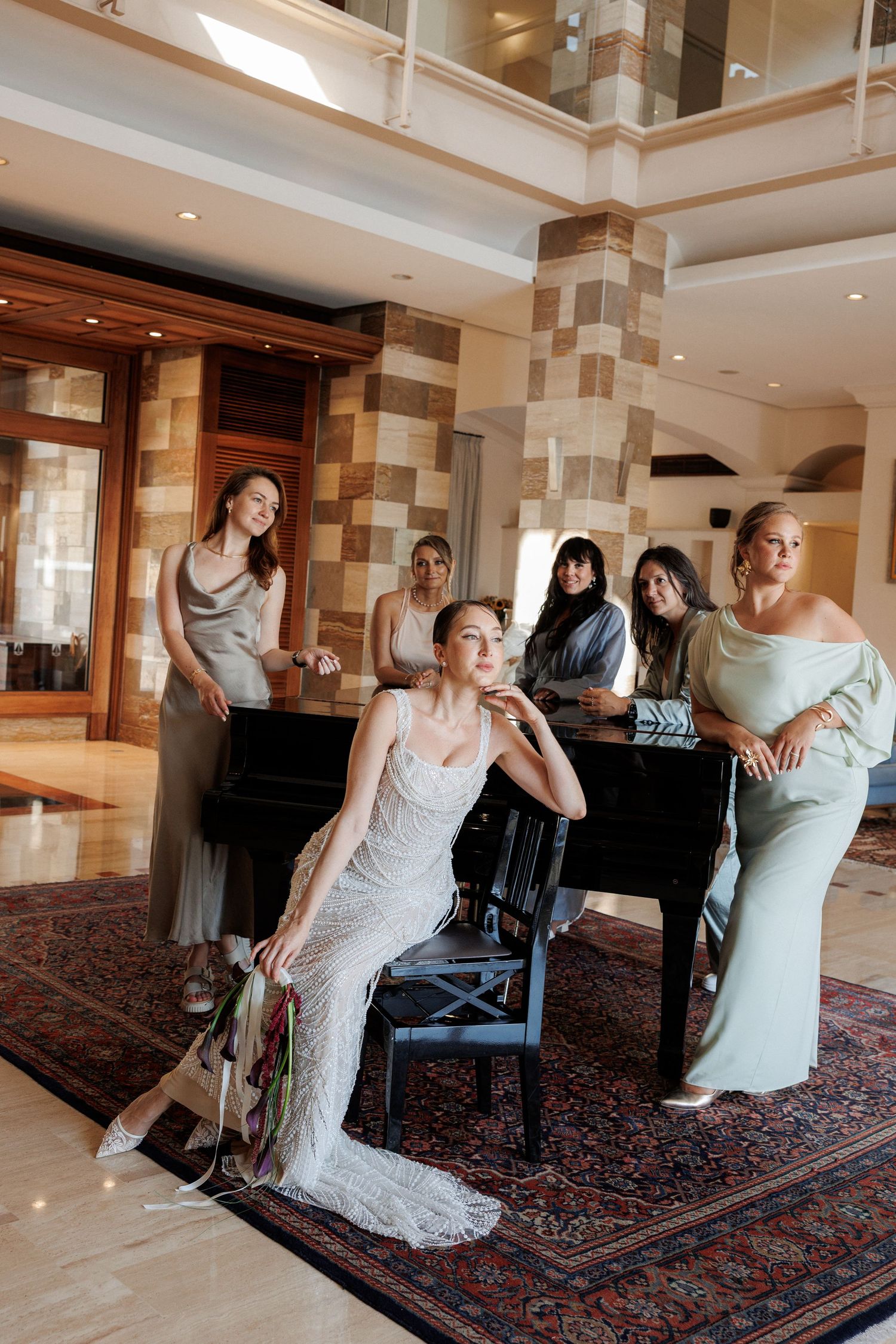 Bride in white gown sits elegantly with bridesmaids in sage green dresses beside a grand piano in a luxurious venue.
