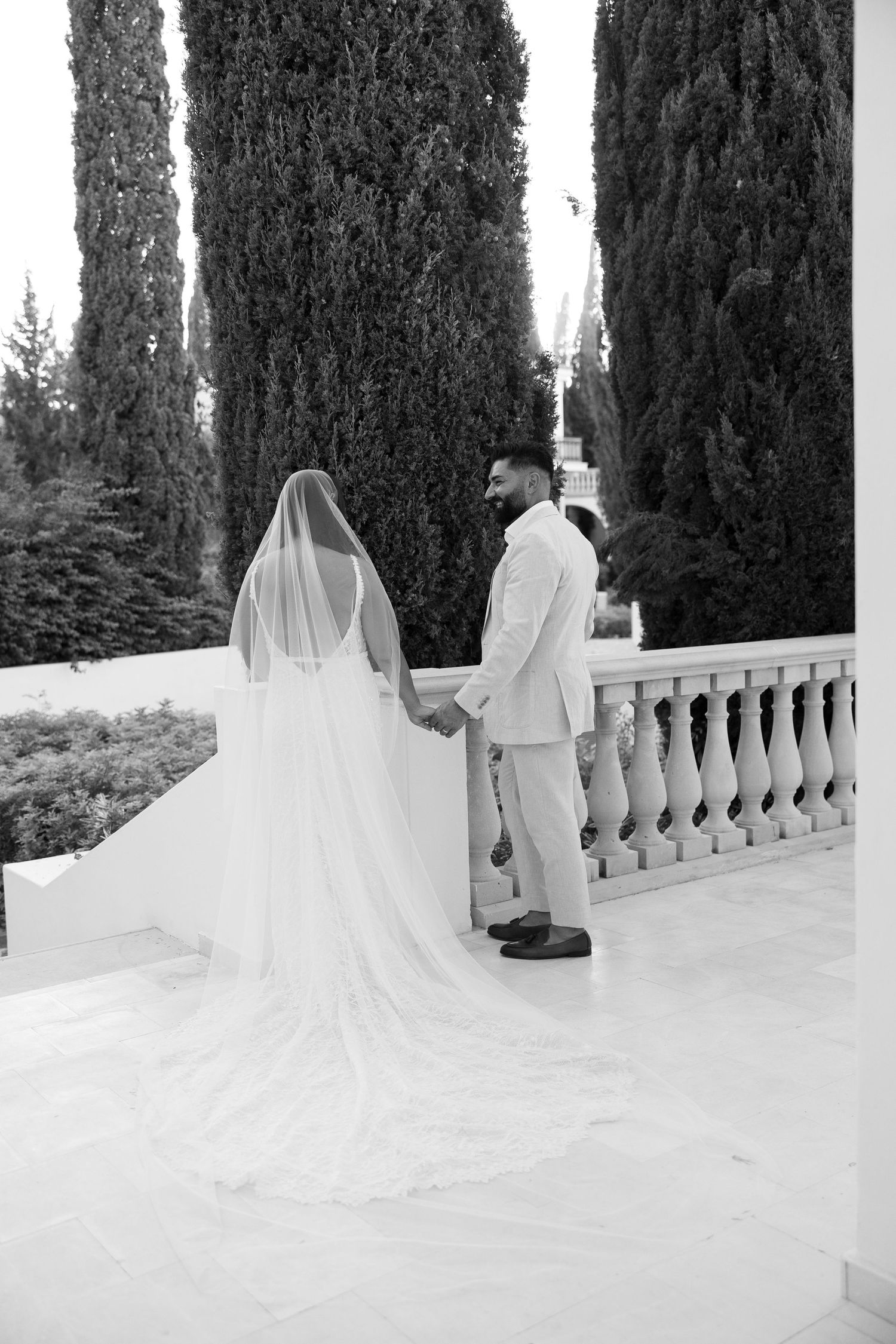 Black and white photo of bride and groom holding hands on a terrace with tall cypress trees.