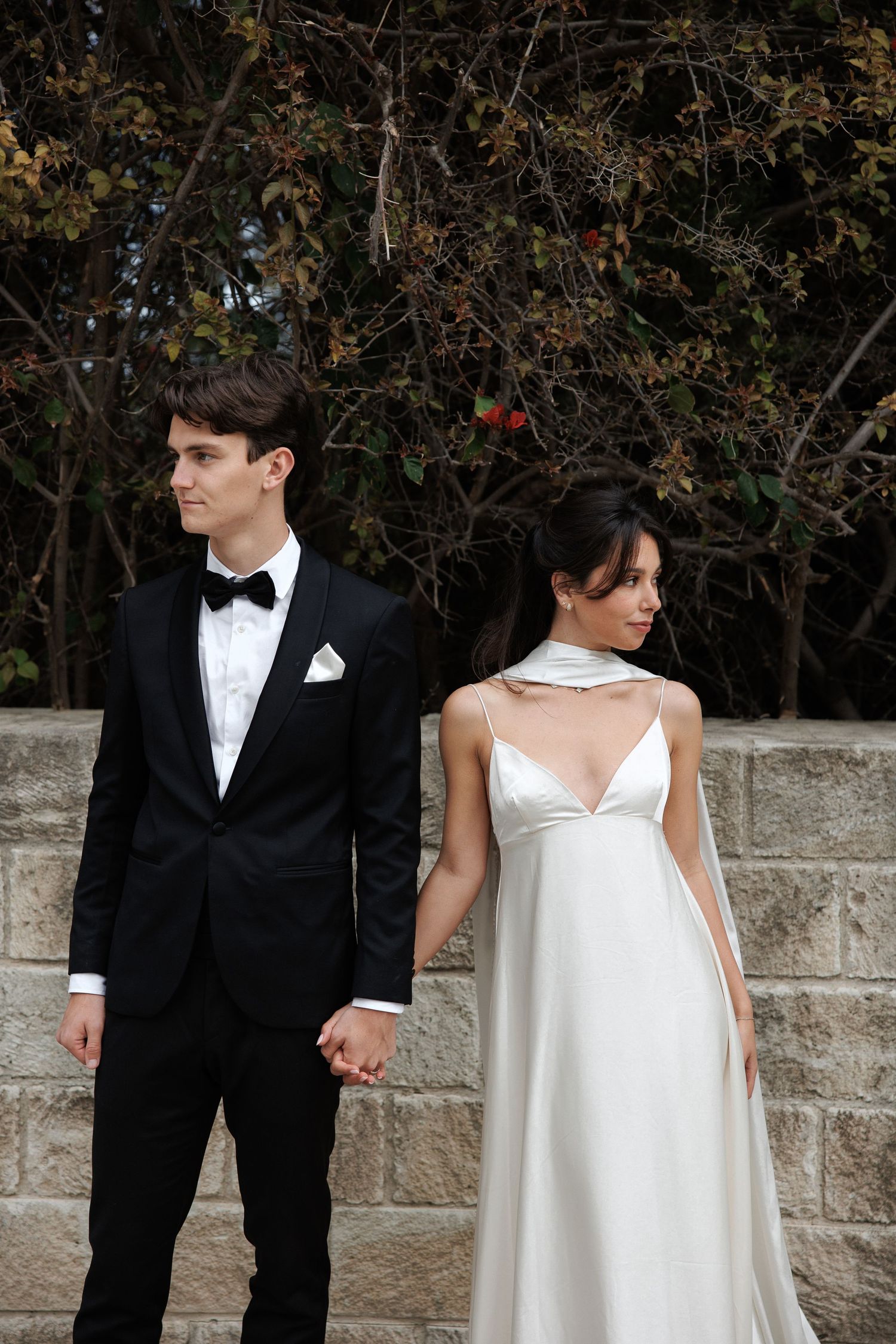 A couple in wedding attire stand against a stone wall while holding hands, dressed in black tuxedo and white gown.