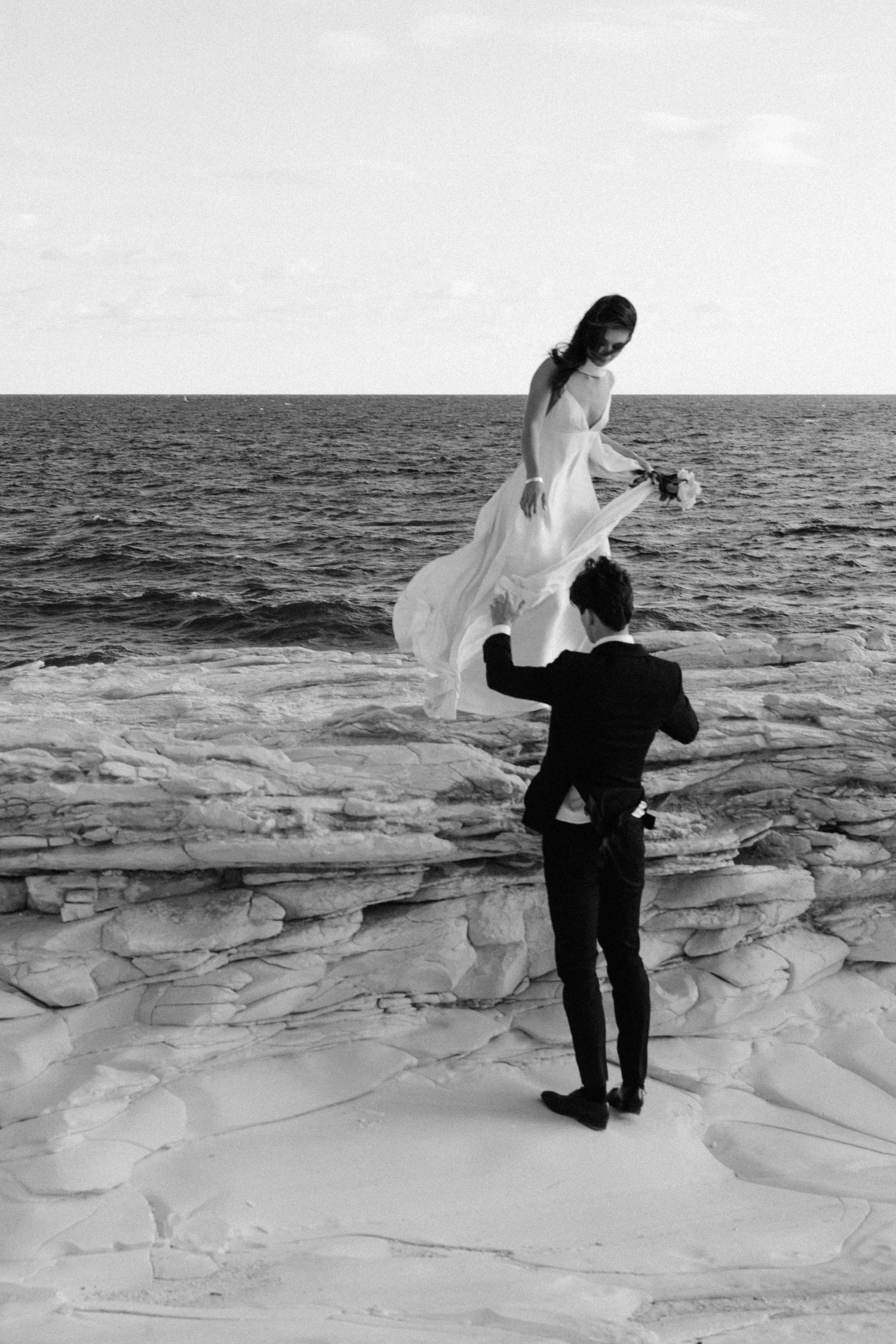 A couple poses for a dramatic black and white wedding photo on coastal rocks by the ocean.