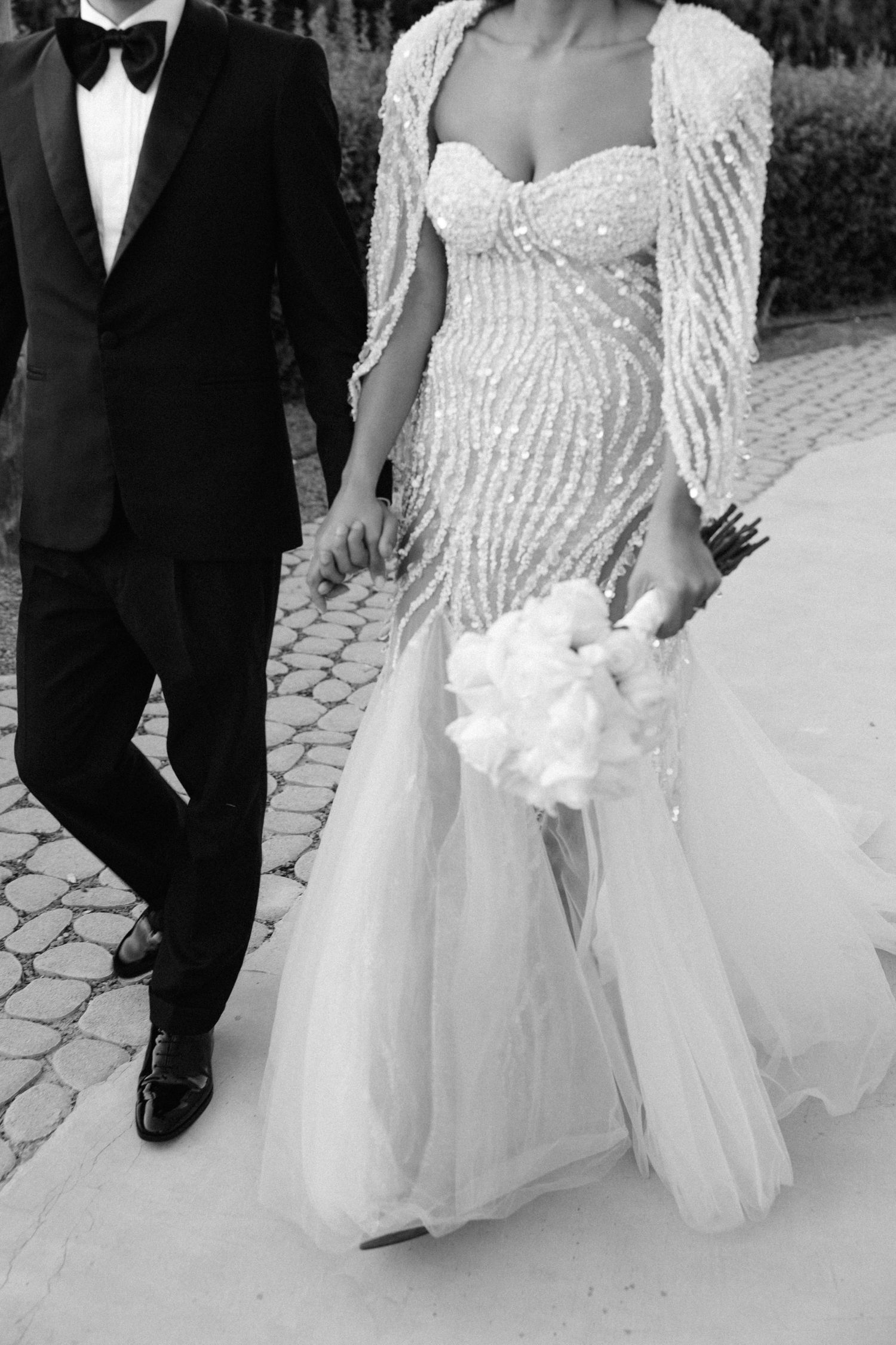 Black and white photo of bride in beaded mermaid gown holding bouquet, walking with groom in tuxedo.