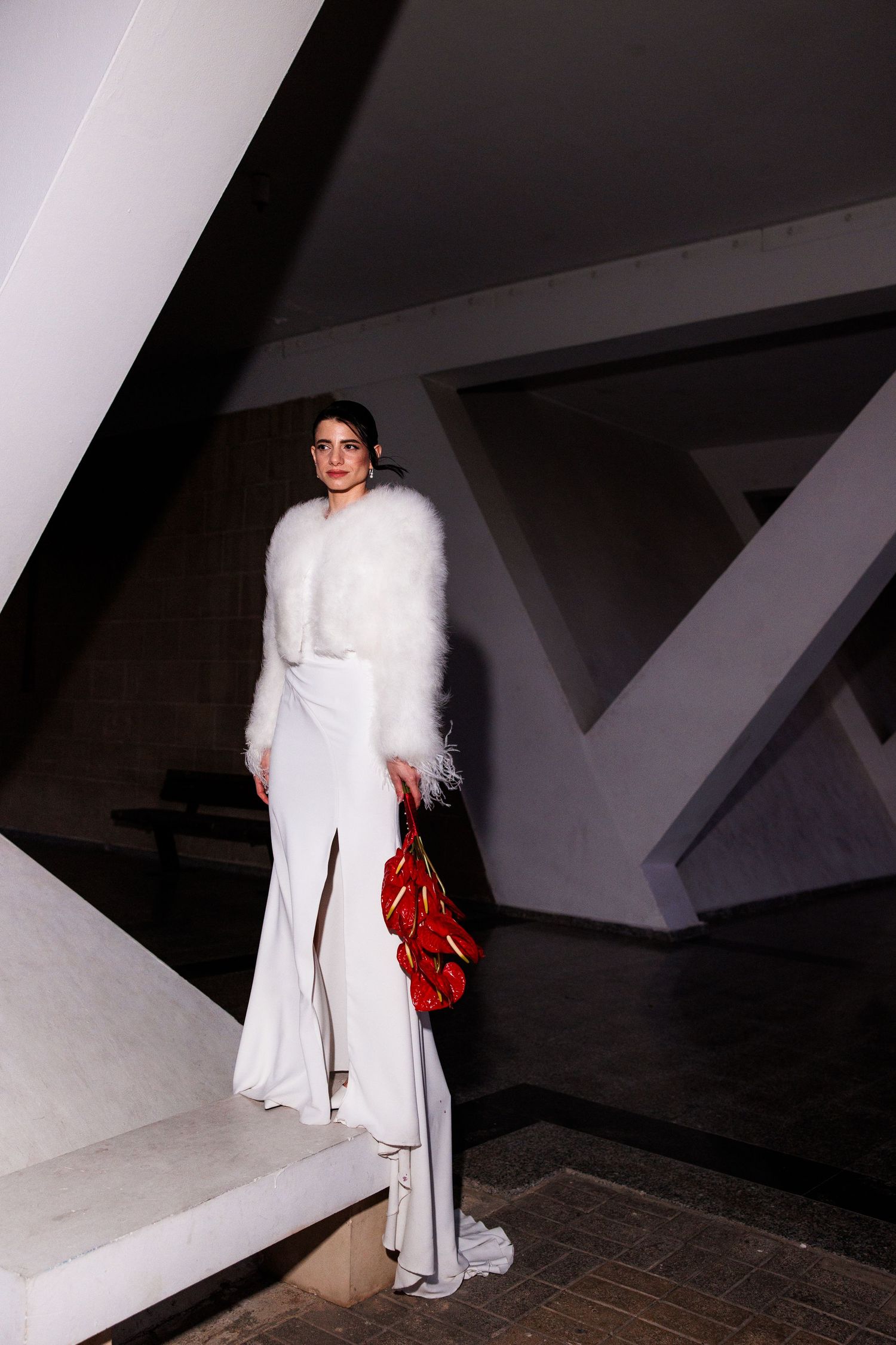 Bride in white gown and fluffy fur jacket holding red floral clutch on modern architectural staircase.