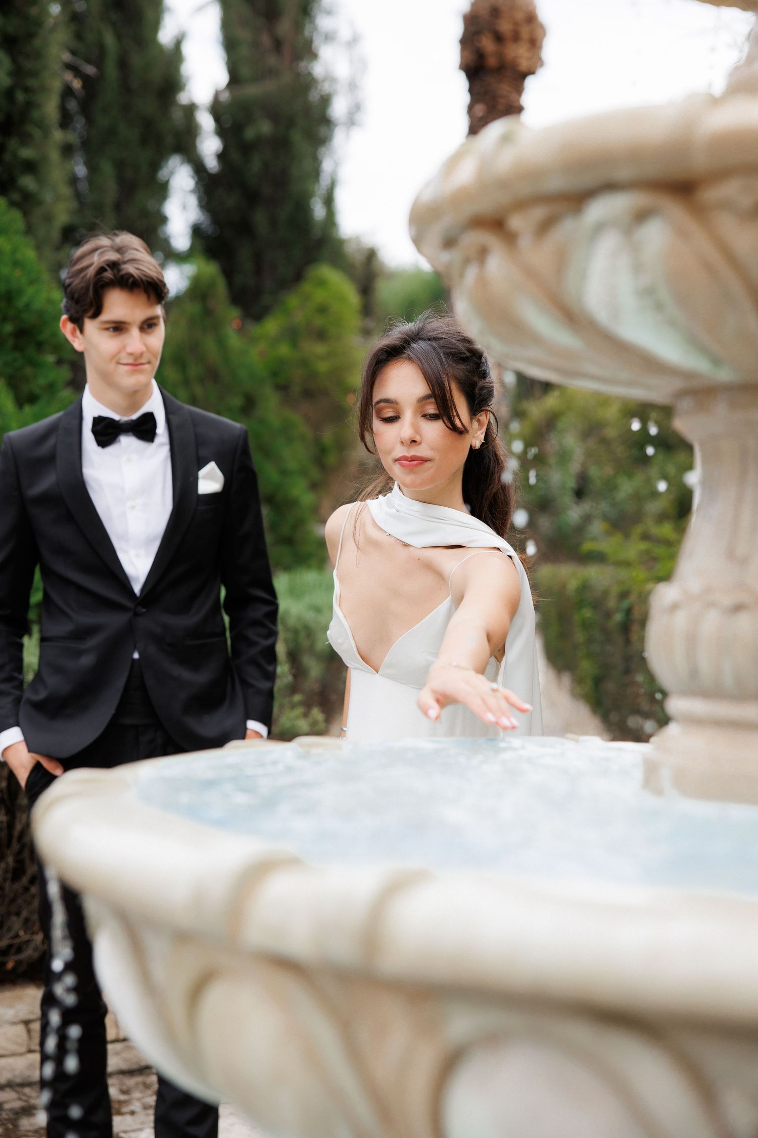 Newlywed couple in formal attire poses near an ornate stone fountain in a garden with cypress trees.