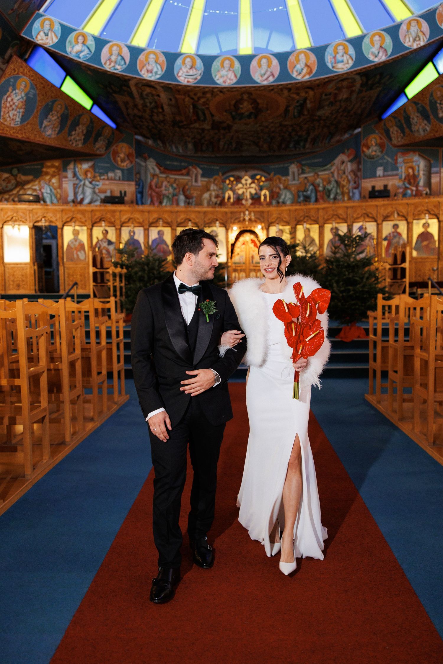 Bride and groom walking down the aisle of an ornate Orthodox church, bride holding red anthurium bouquet.