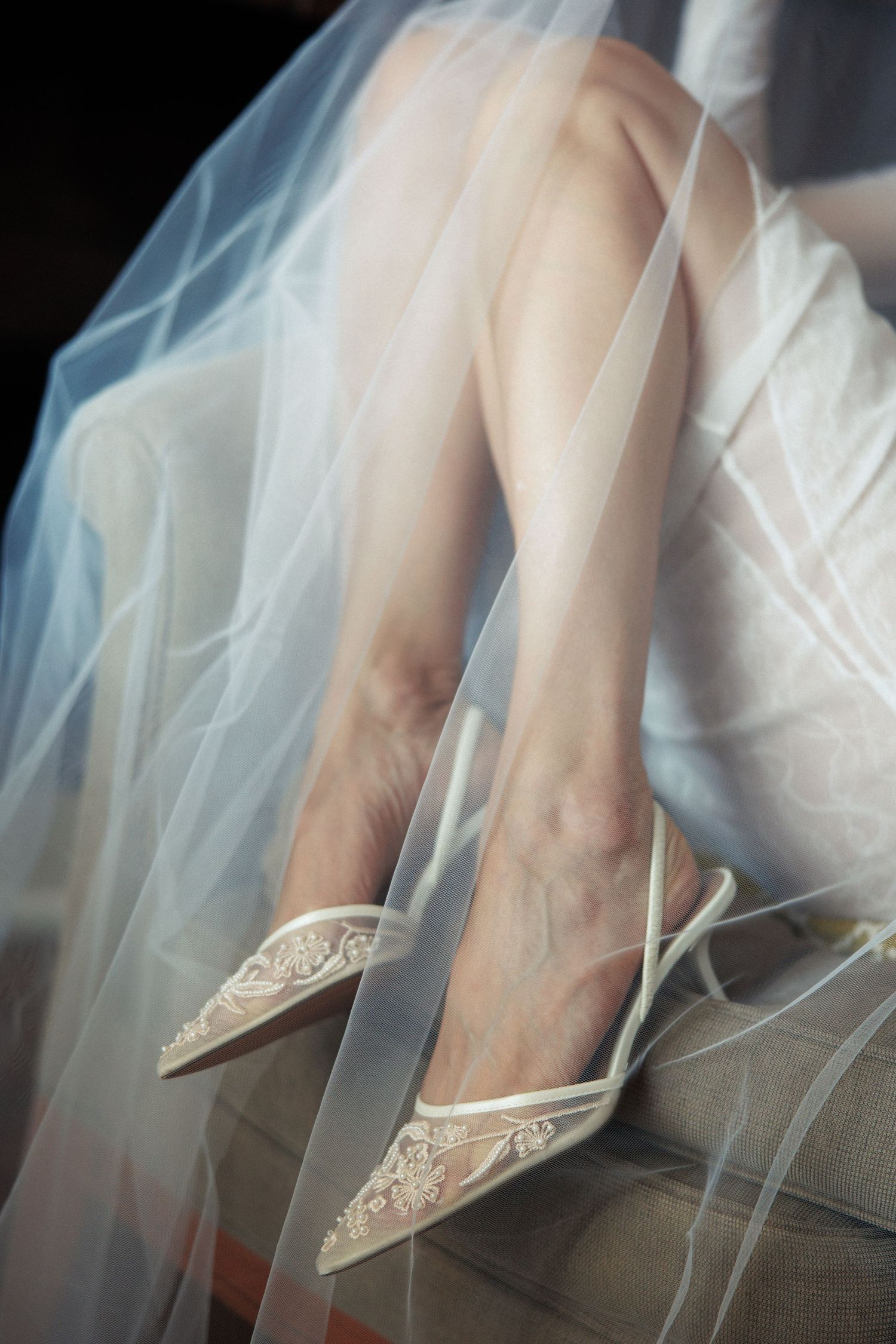 Bride wearing elegant white lace pointed-toe heels with delicate straps beneath a flowing tulle wedding veil.