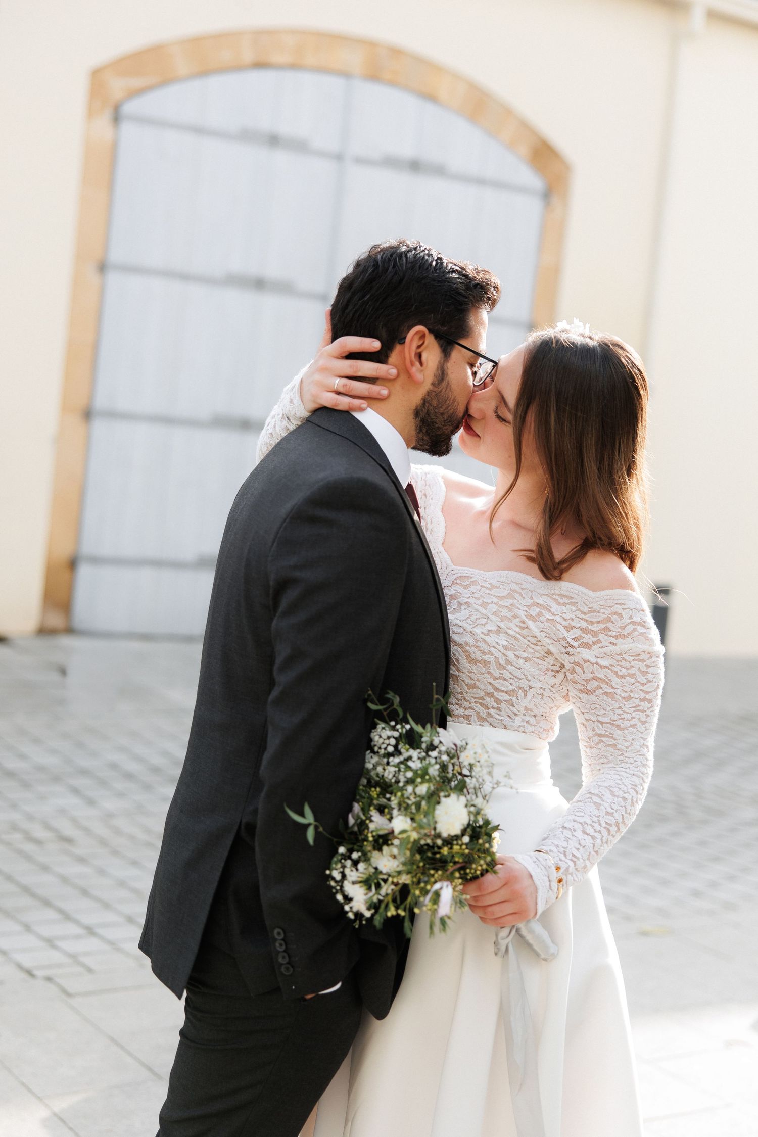 Bride and groom sharing a romantic kiss outdoors, bride holding white floral bouquet in lace long-sleeve wedding dress.