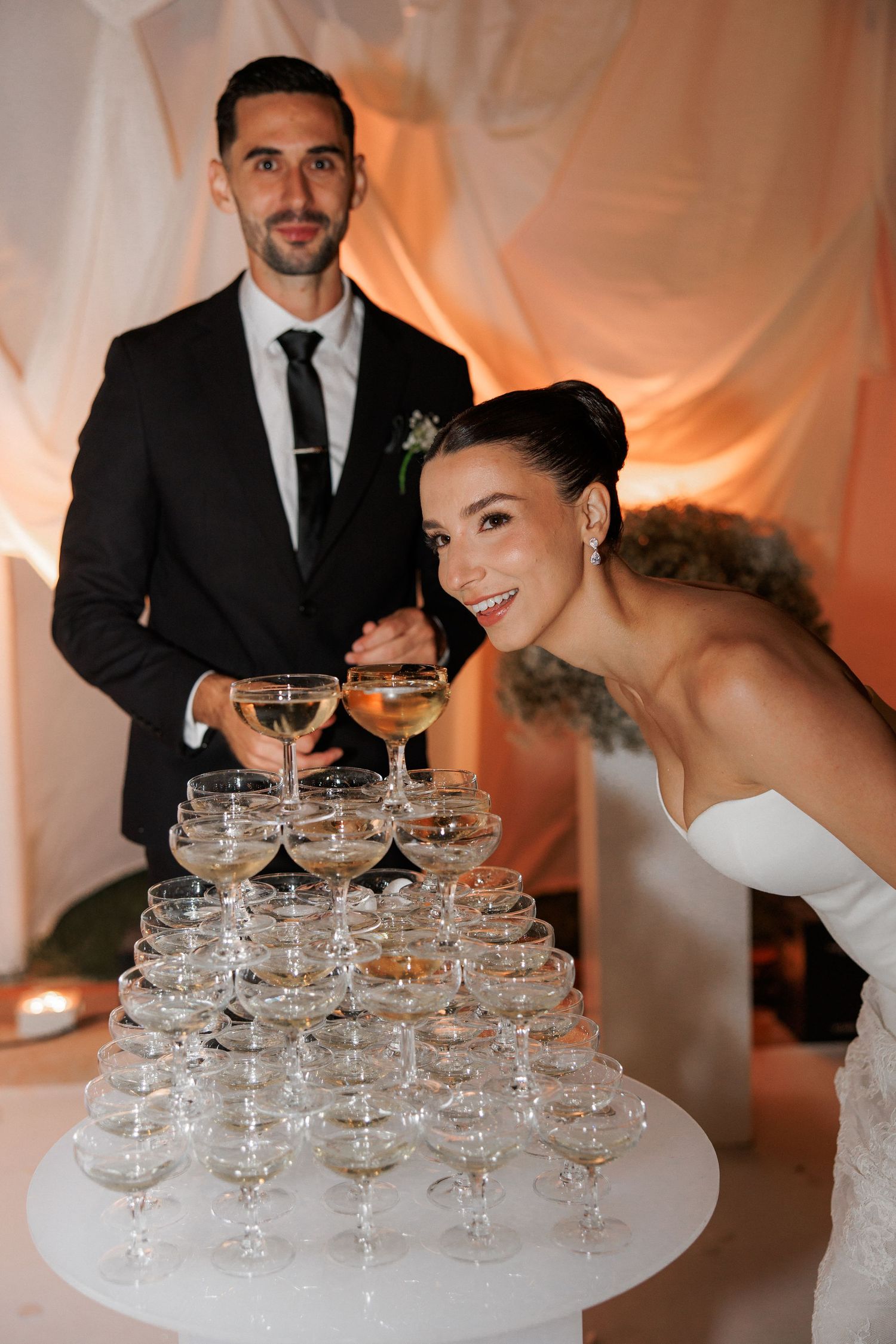 Wedding couple posing next to a tower of champagne glasses at their reception.