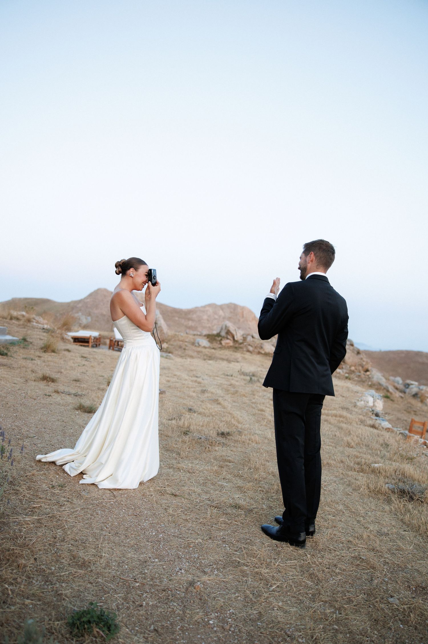 Bride and groom photograph each other on scenic desert hillside during golden hour wedding shoot.