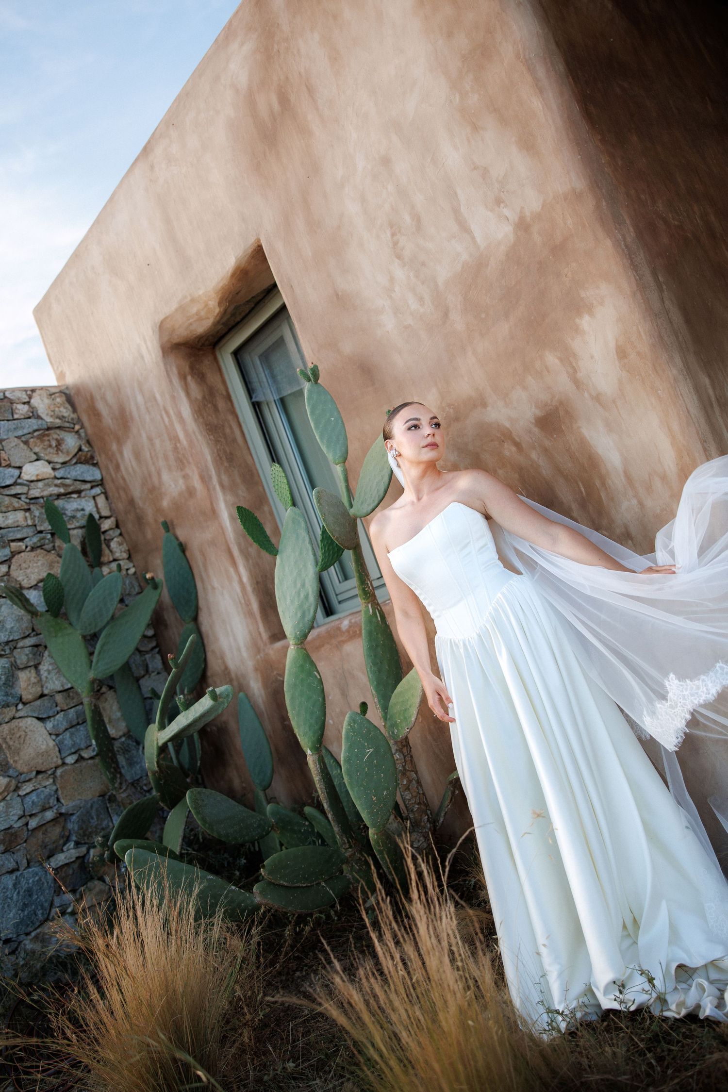 Bride in white strapless gown with flowing veil poses near cactus by rustic adobe wall.