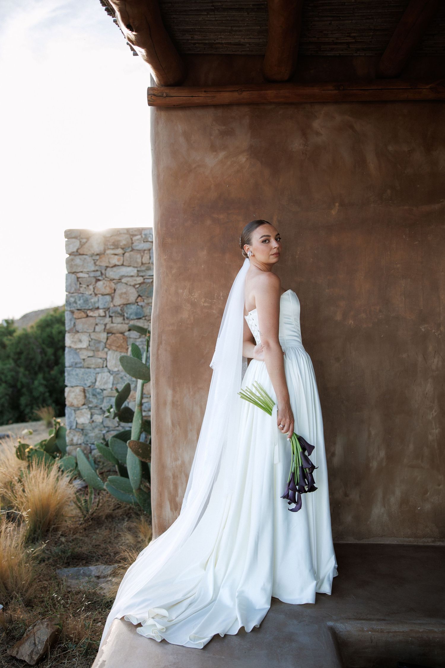 Bride in white strapless gown with veil holds bouquet beside rustic adobe wall in desert setting.