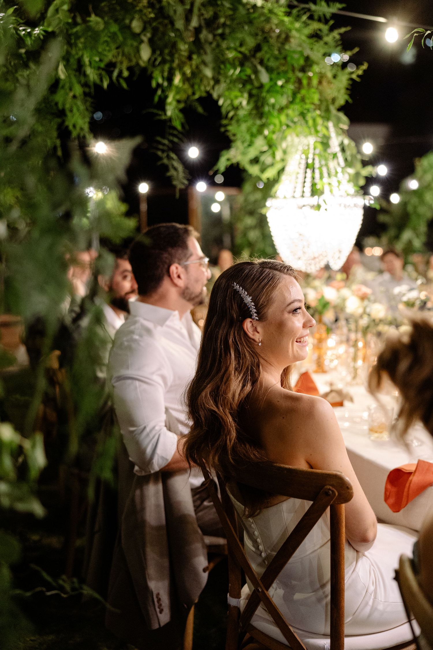 Romantic evening wedding reception under crystal chandeliers and twinkling lights with greenery overhead.