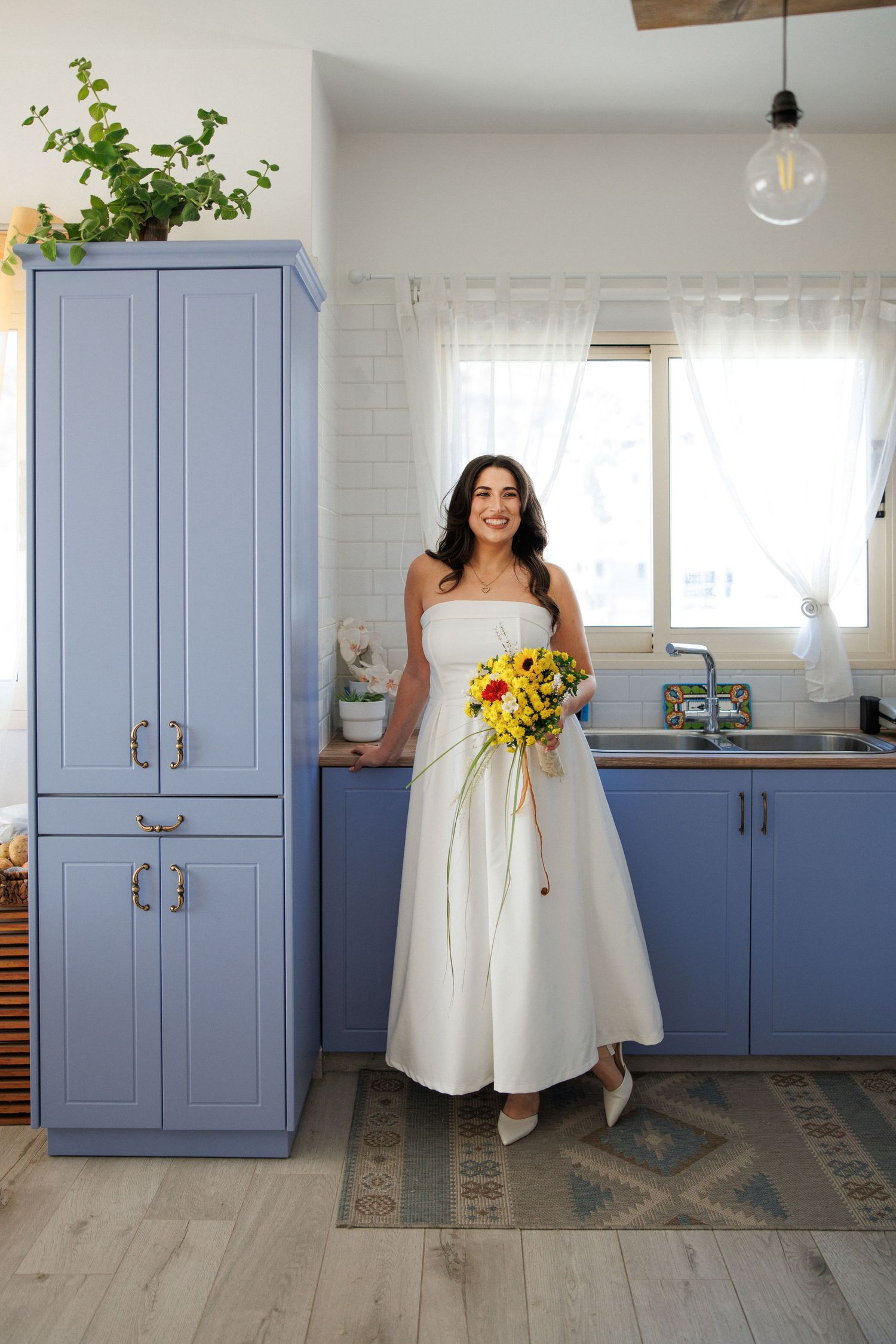 A woman in a white wedding dress holds a sunflower bouquet in a kitchen with blue cabinets.