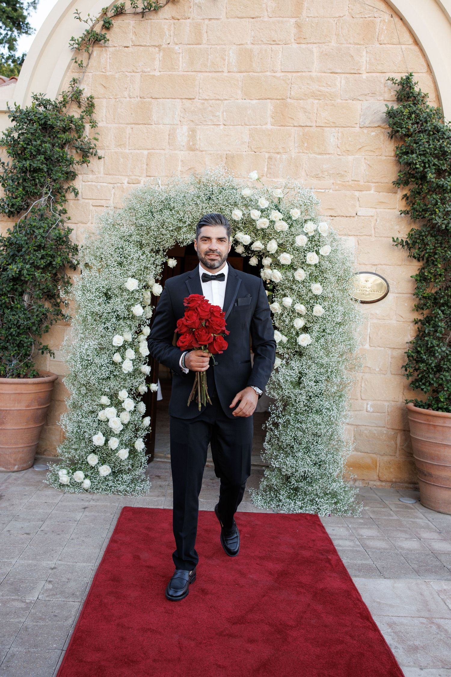 Groom in black tuxedo holding red roses bouquet on red carpet before floral arch at wedding venue.