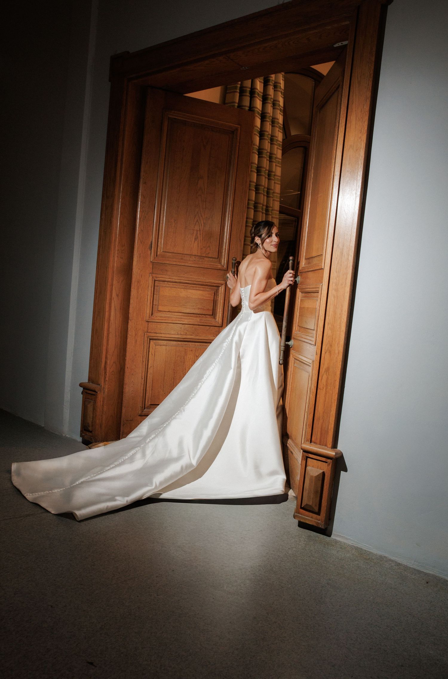 Bride in elegant white satin ball gown with long train opening ornate wooden door in grand interior.