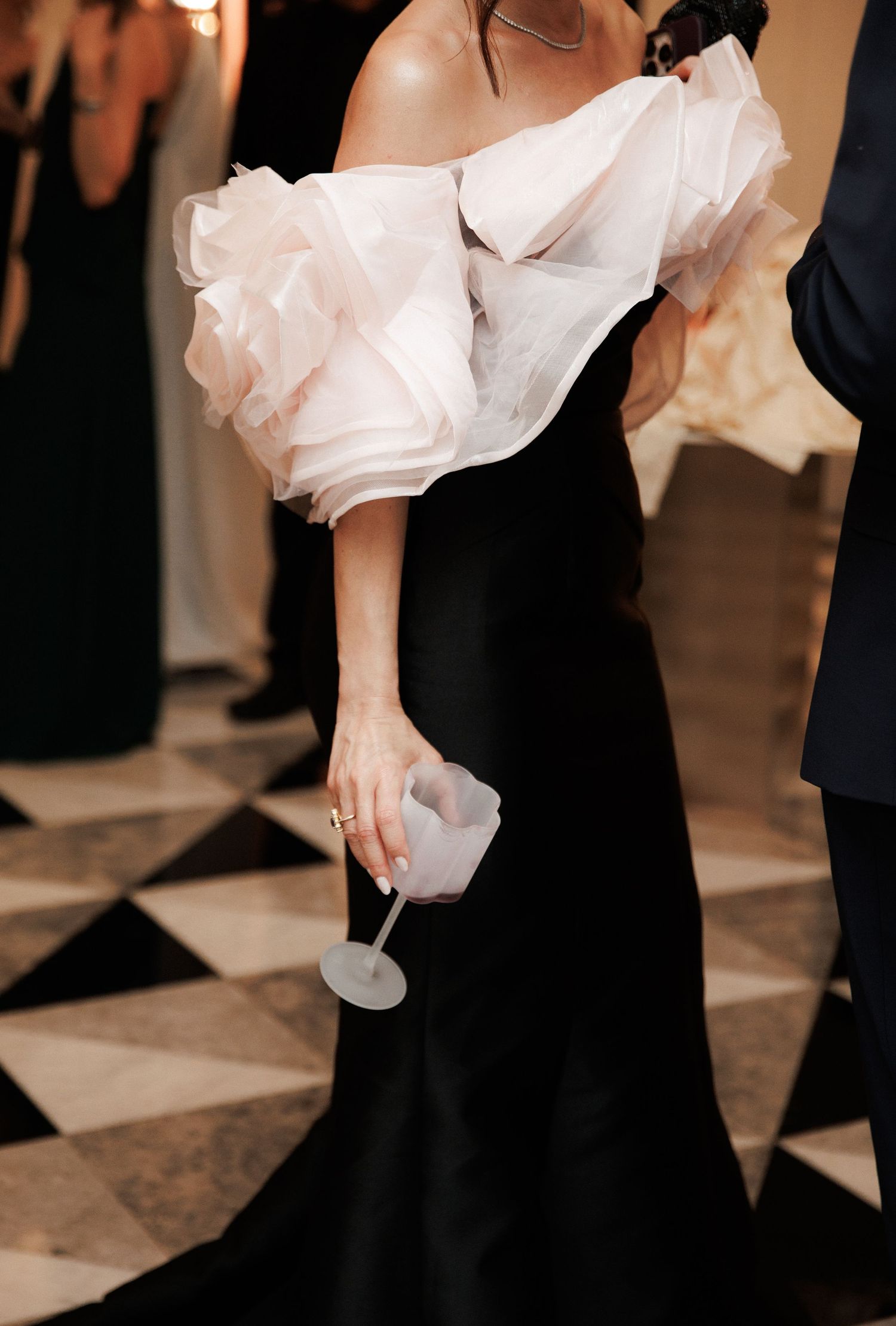 Elegant woman in black gown with white ruffled off-shoulder top holding a cocktail glass at formal event.