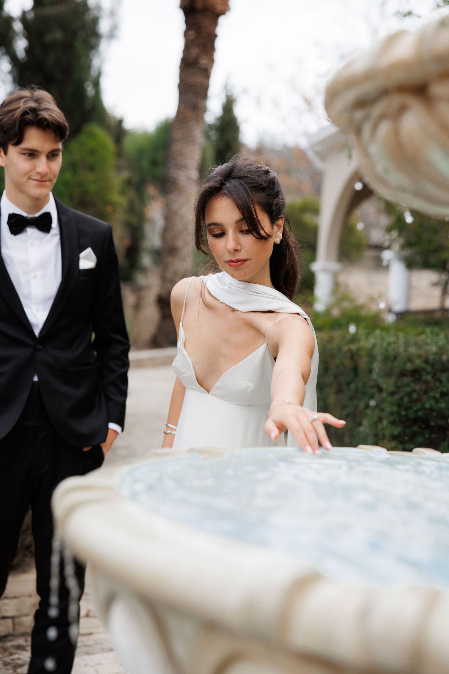 Elegant couple in formal attire by a decorative fountain in a garden setting.