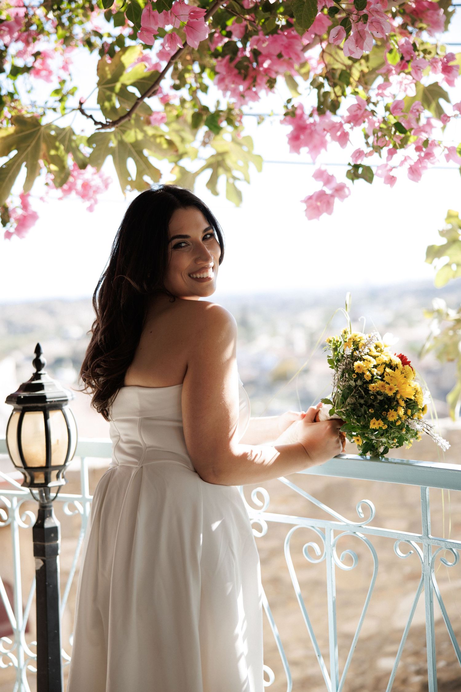 A bride poses with her bouquet on a balcony under pink flowering vines with a scenic backdrop.
