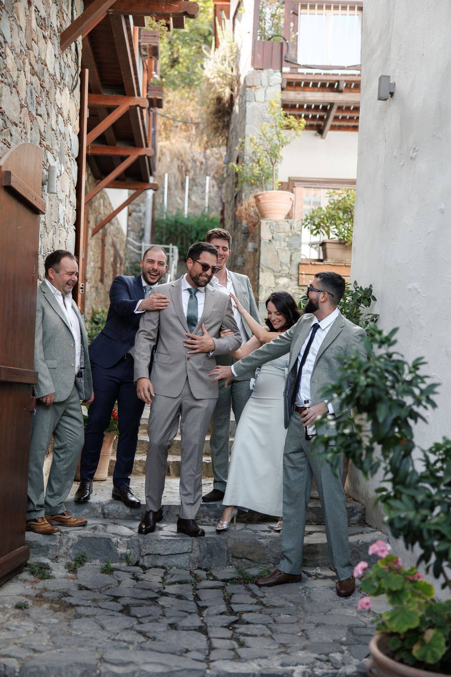 Wedding party in formal attire gathered on a charming cobblestone alley between rustic stone buildings.