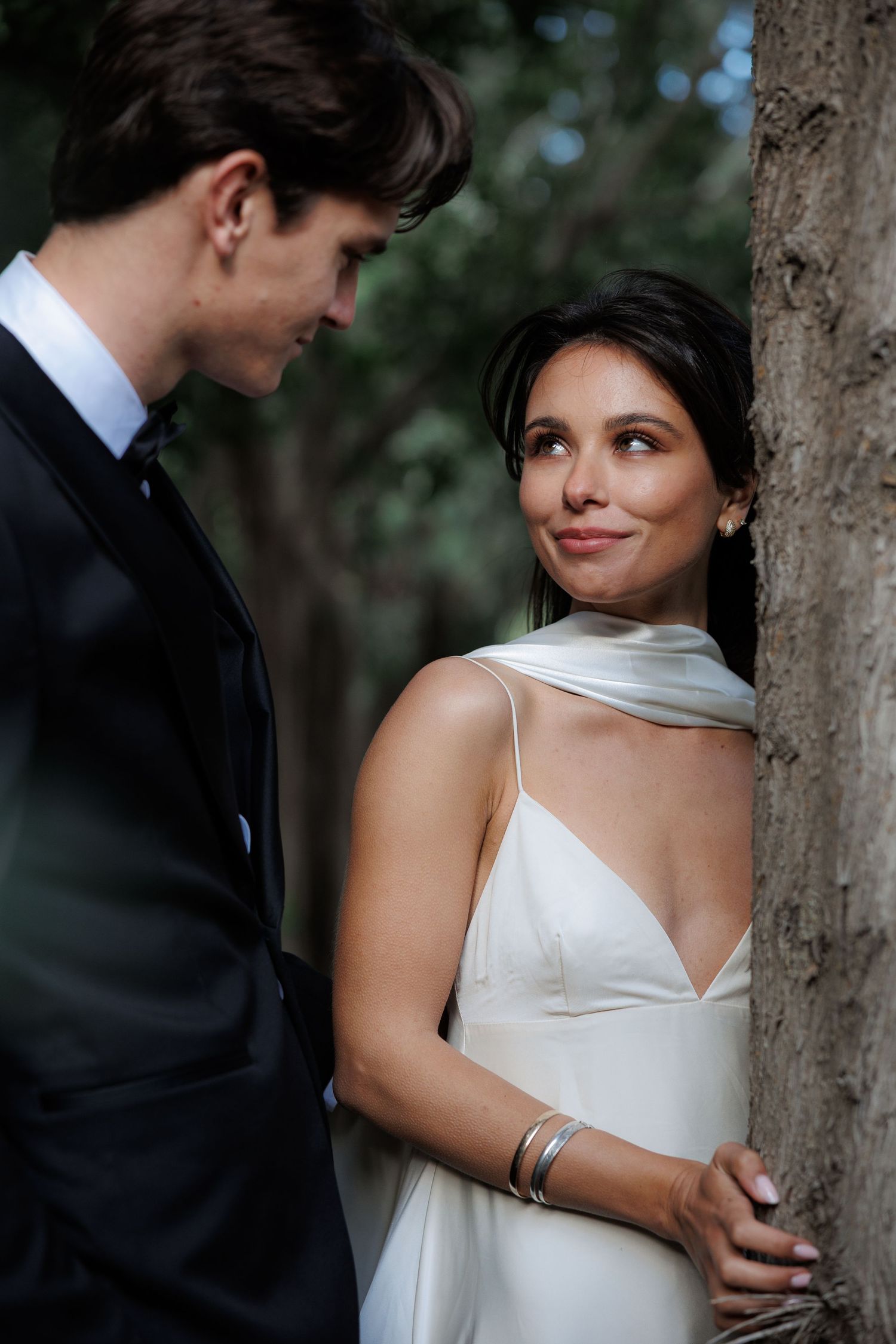 A couple in formal wedding attire sharing an intimate moment next to a tree in a romantic outdoor setting.