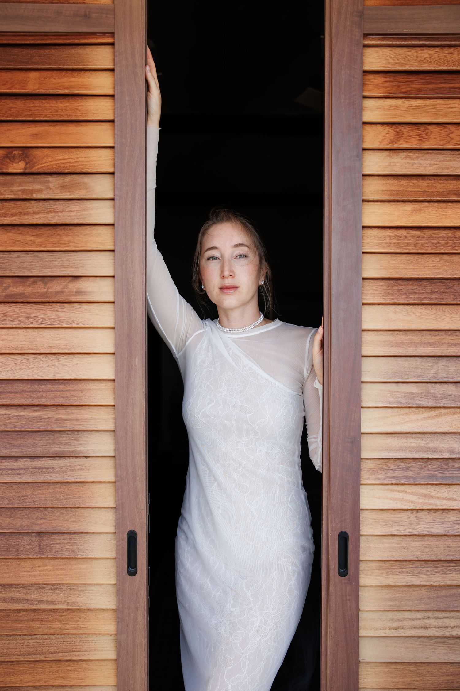 Woman in white lace dress standing in wooden doorway with arm raised, elegant bridal portrait.