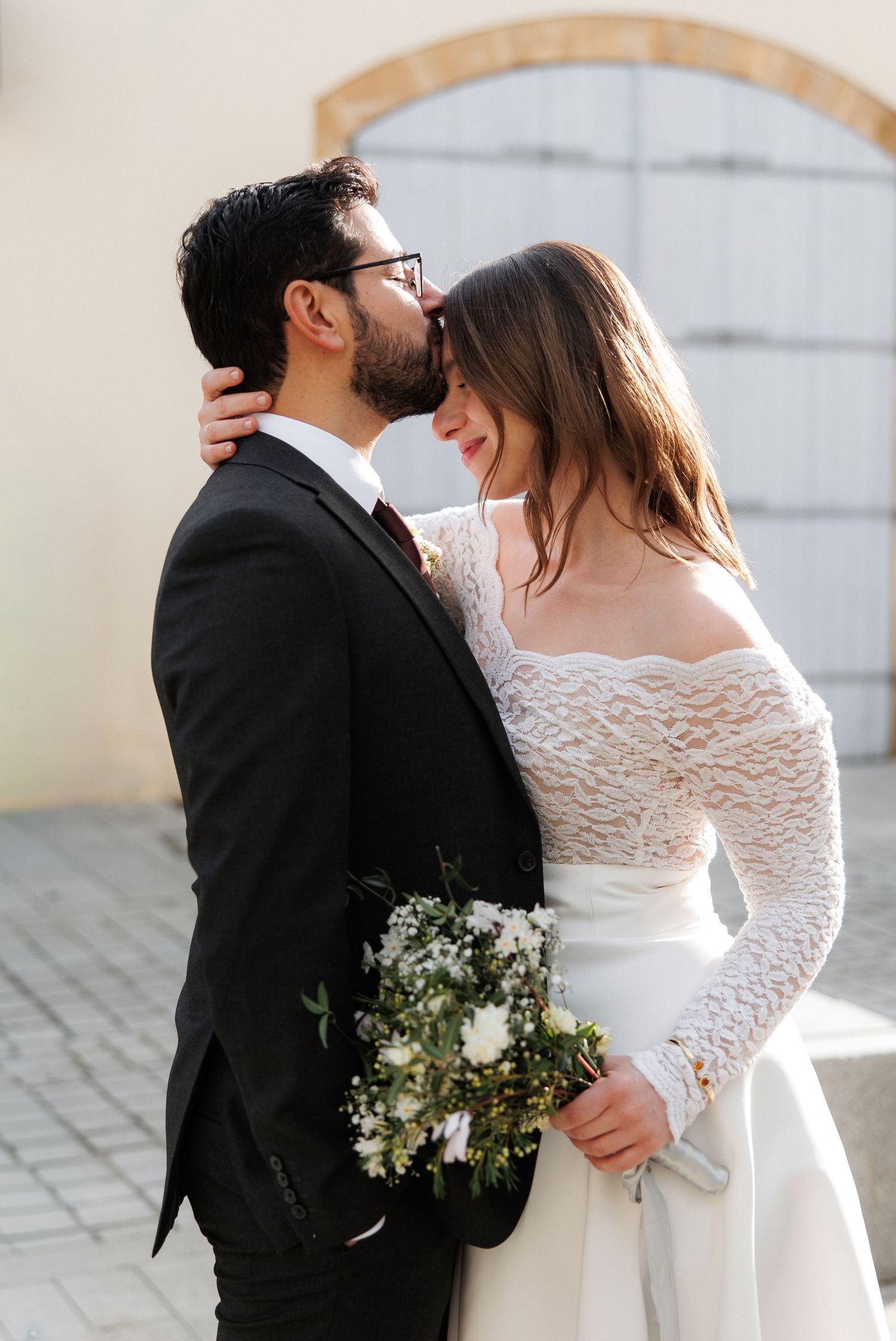 Bride and groom share a tender moment, she holds white floral bouquet, wearing lace off-shoulder gown, he in black suit.